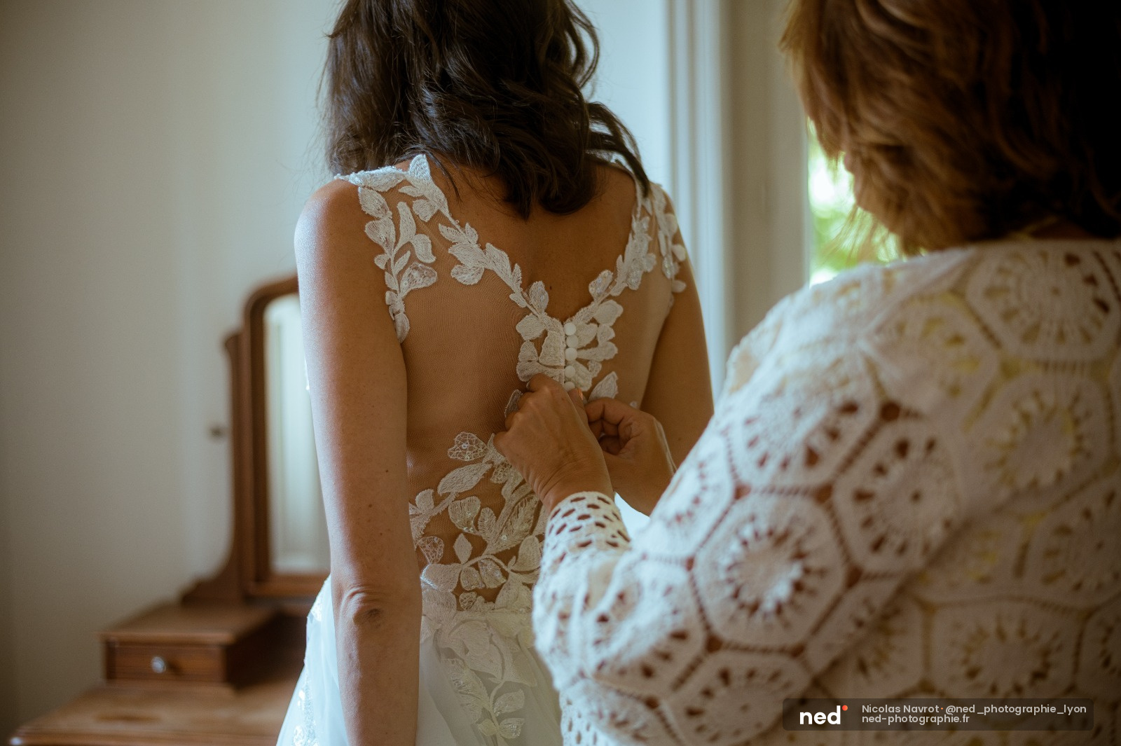 Mariée de dos en robe blanche dentelle tandis qu'une femme ajuste les lacets, lumière naturelle dans la chambre