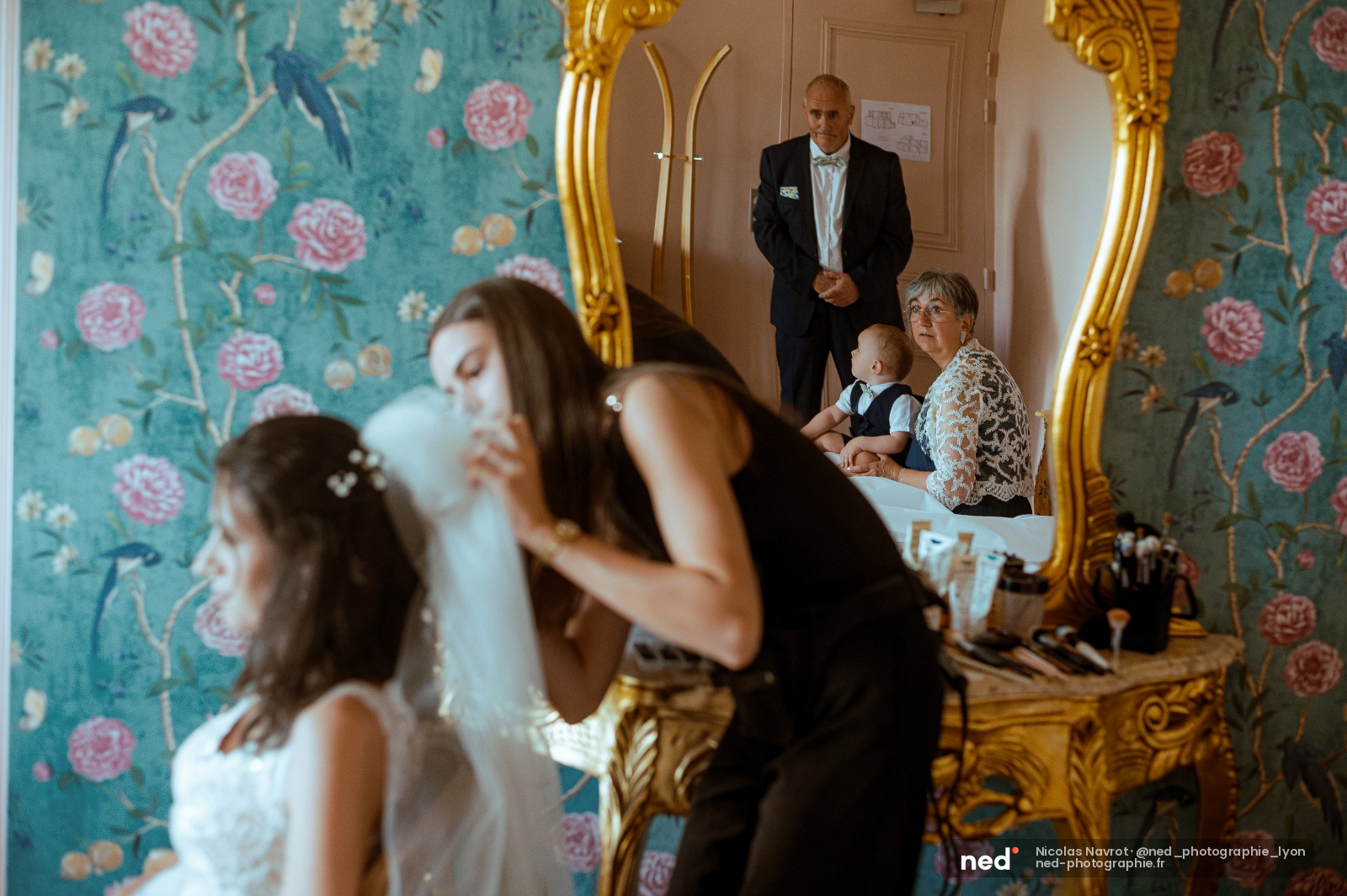 Mariée en robe blanche ajuste son voile devant un grand miroir doré, famille et enfant observent dans une pièce aux murs bleu turquoise à motifs floraux