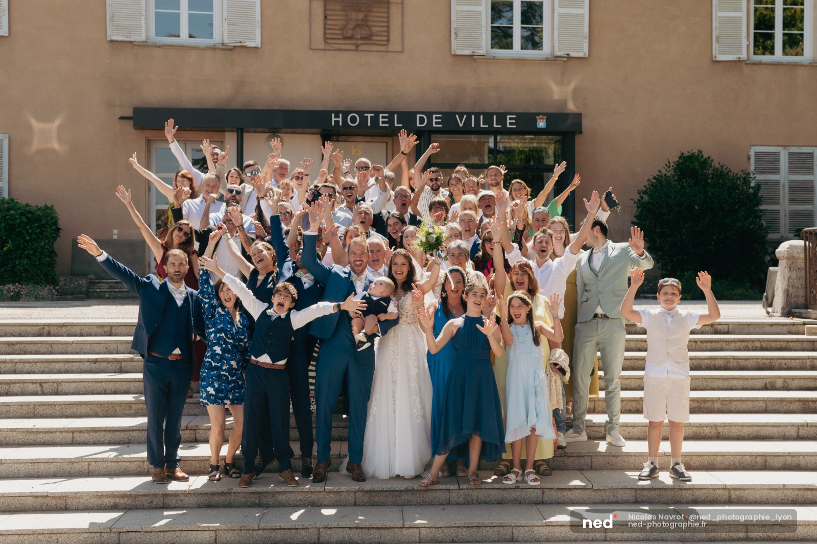 Cortege de mariage de plus de 50 personnes, bras leves, devant l'Hotel de Ville avec facade beige et volets blancs, soleil de midi