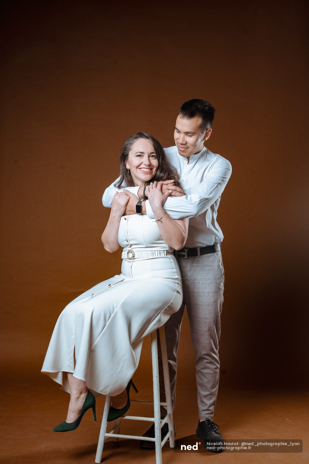 Femme en robe blanche assise sur un tabouret, homme en chemise bleue debout derrière elle, sourires complices, fond or.
