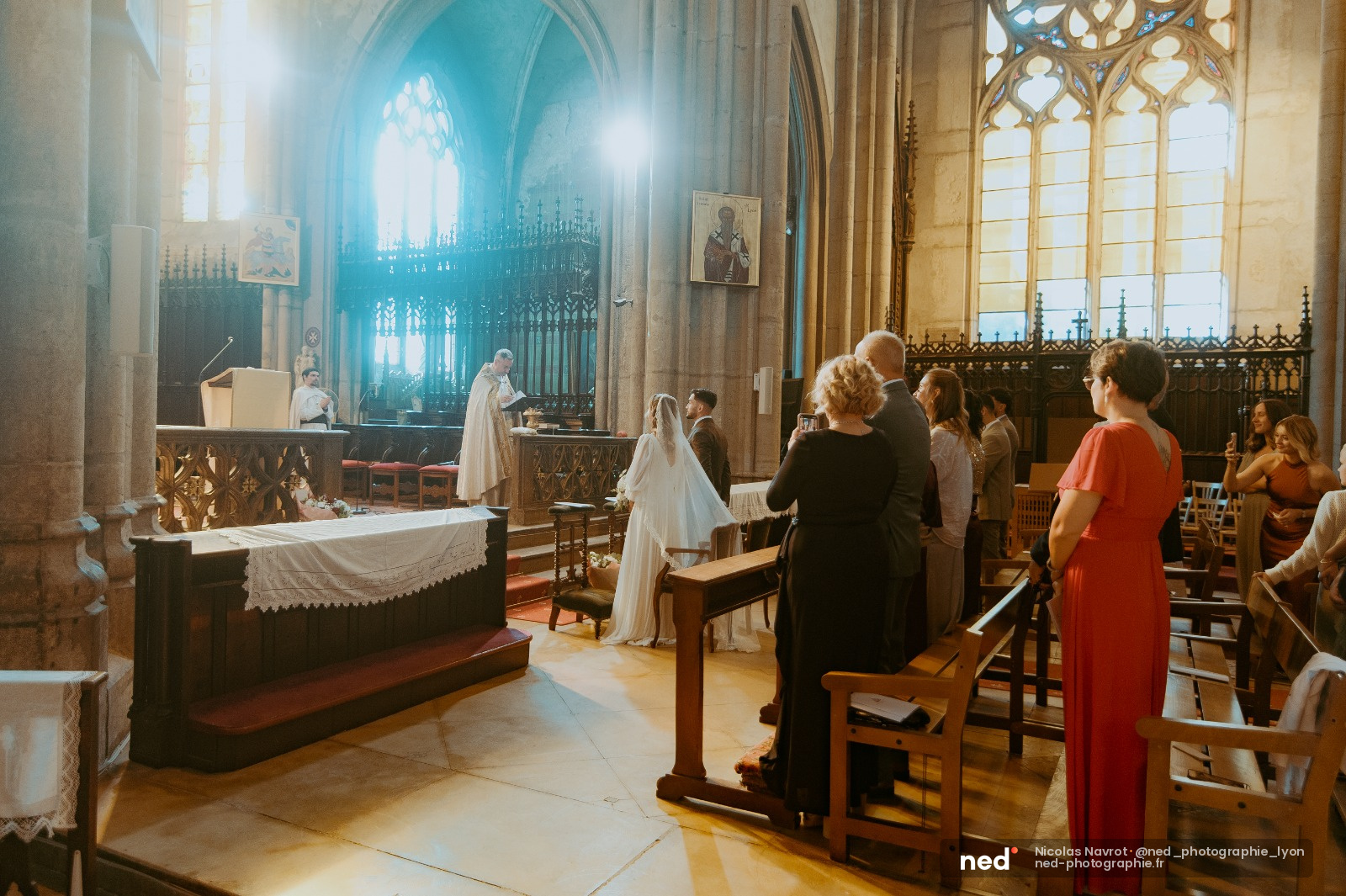 Mariée en robe blanche et voile debout à l'autel dans une église gothique, entourée d'invités assis, lumière du jour traversant les vitraux