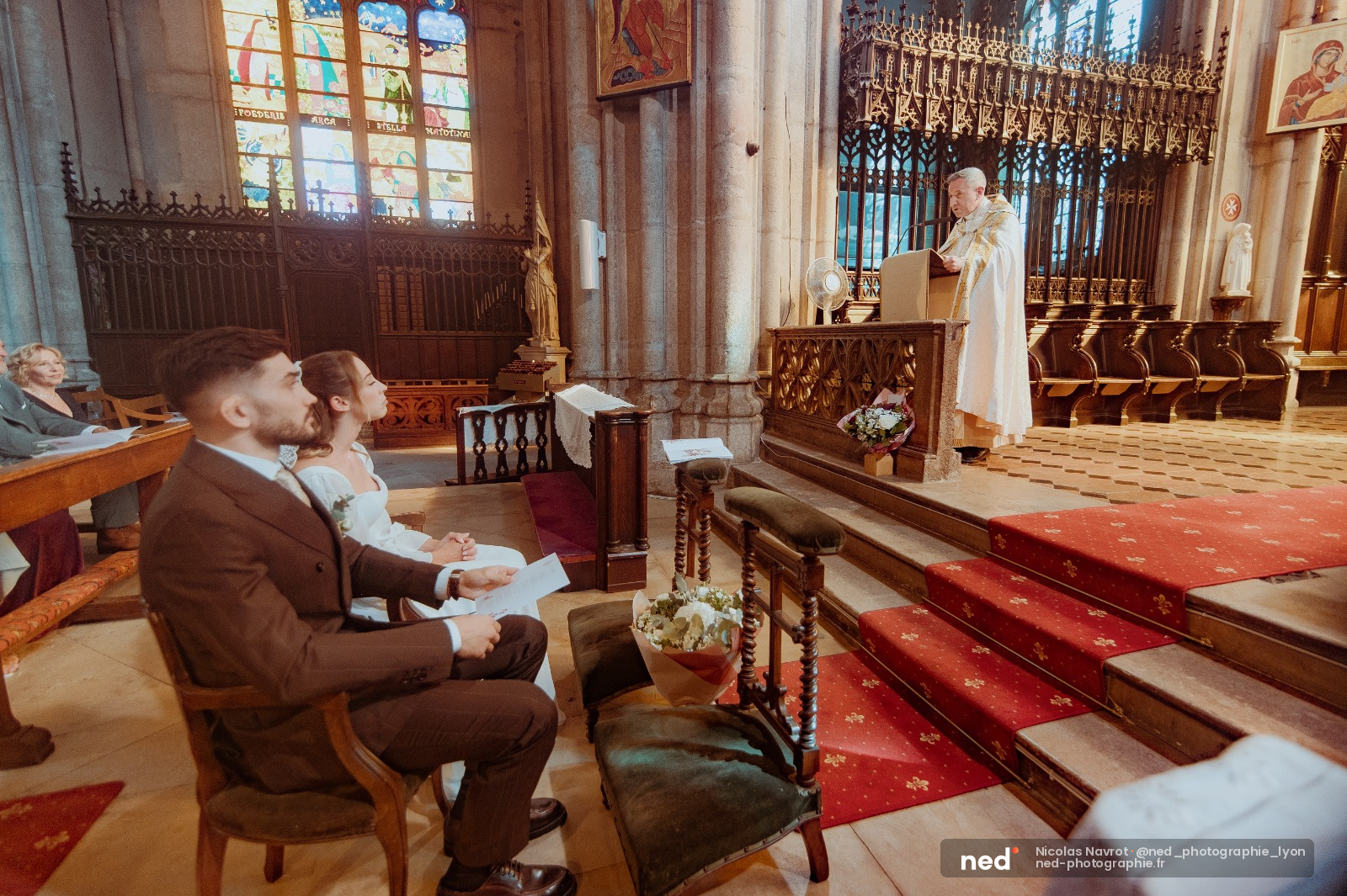 Mariés en costume et robe blanche assis dans les bancs d'une église, prêtre à l'autel, lumière naturelle par les vitraux