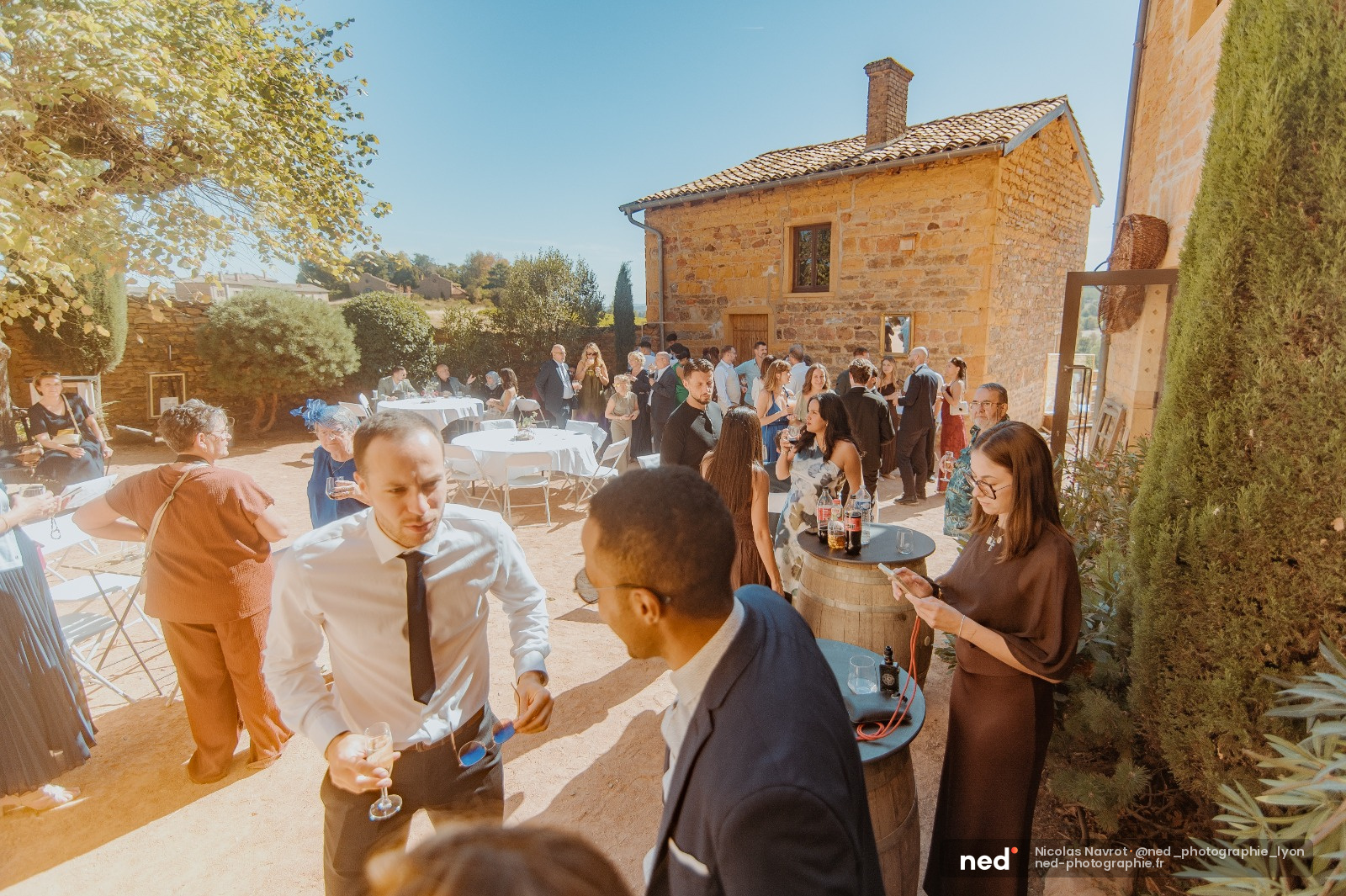 Invités en costume et robe discutent lors d'un cocktail de mariage dans une cour en pierre, sous le soleil, avec tables blanches et bastide en arrière-plan