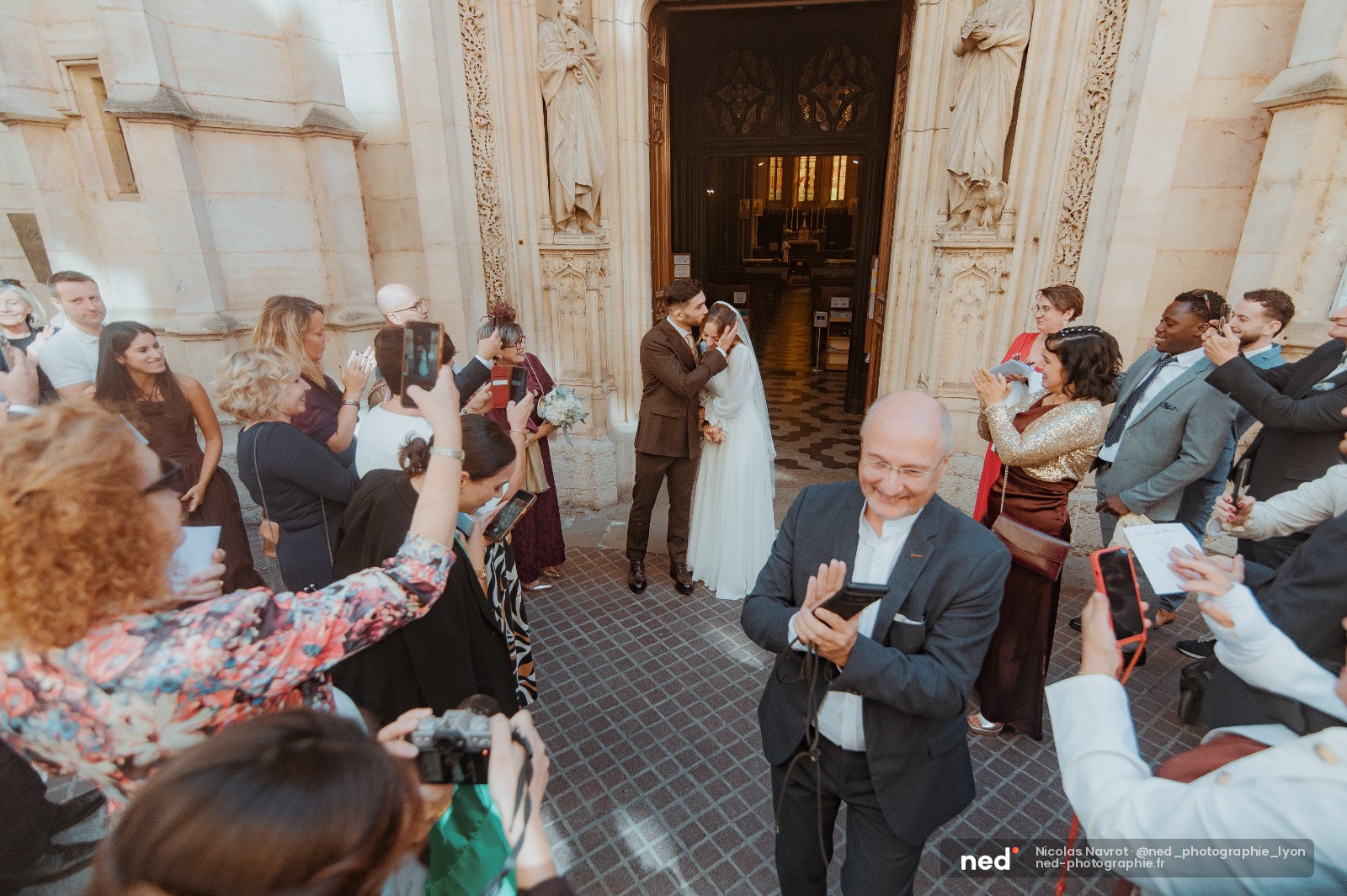 Couple en tenue de mariage s'embrasse au centre d'une allée d'église, invités autour applaudissent et photographient la scène