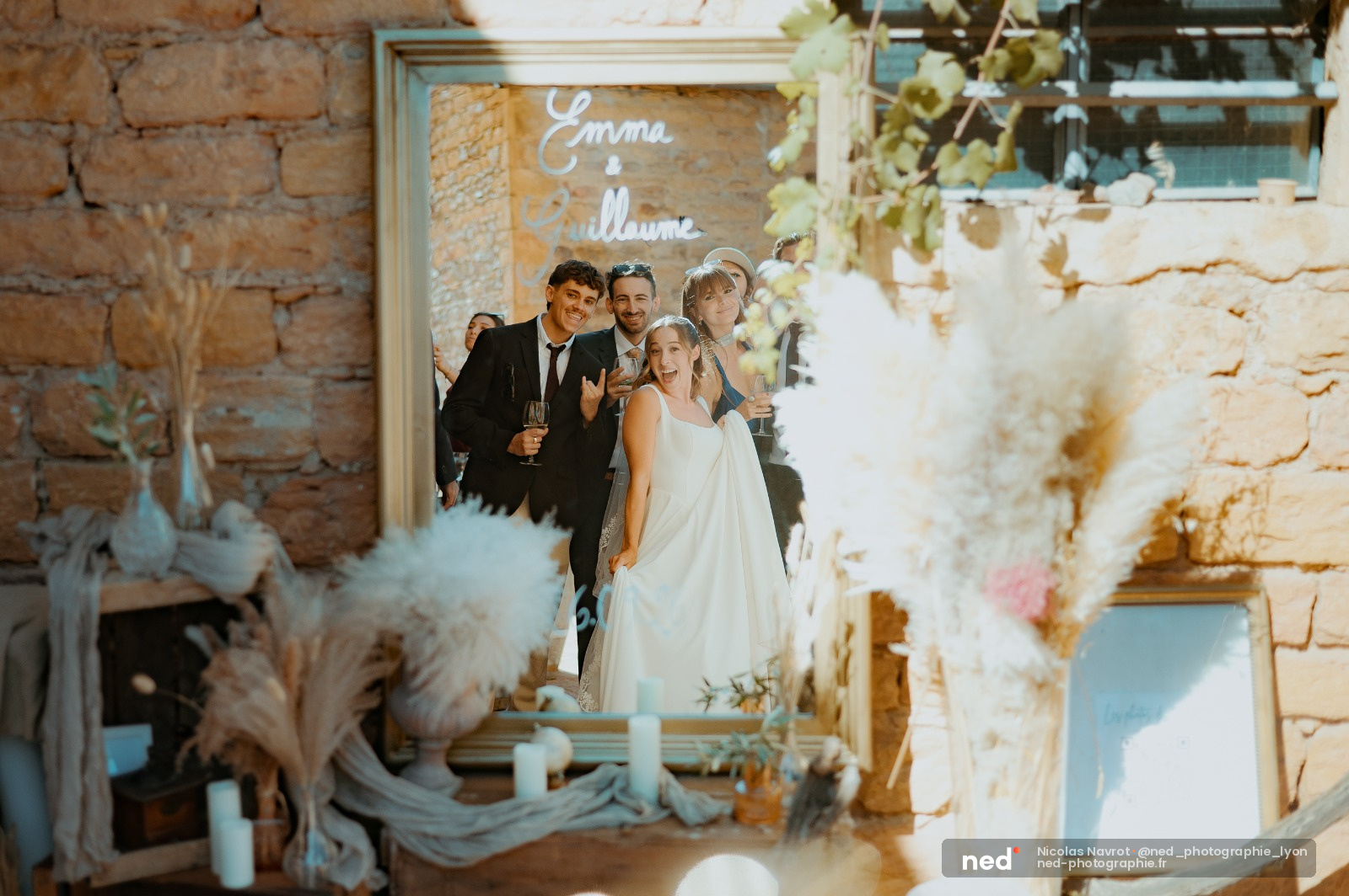Mariée en robe blanche et marié en costume noir posent avec des invités devant un miroir doré dans une salle aux murs en pierre