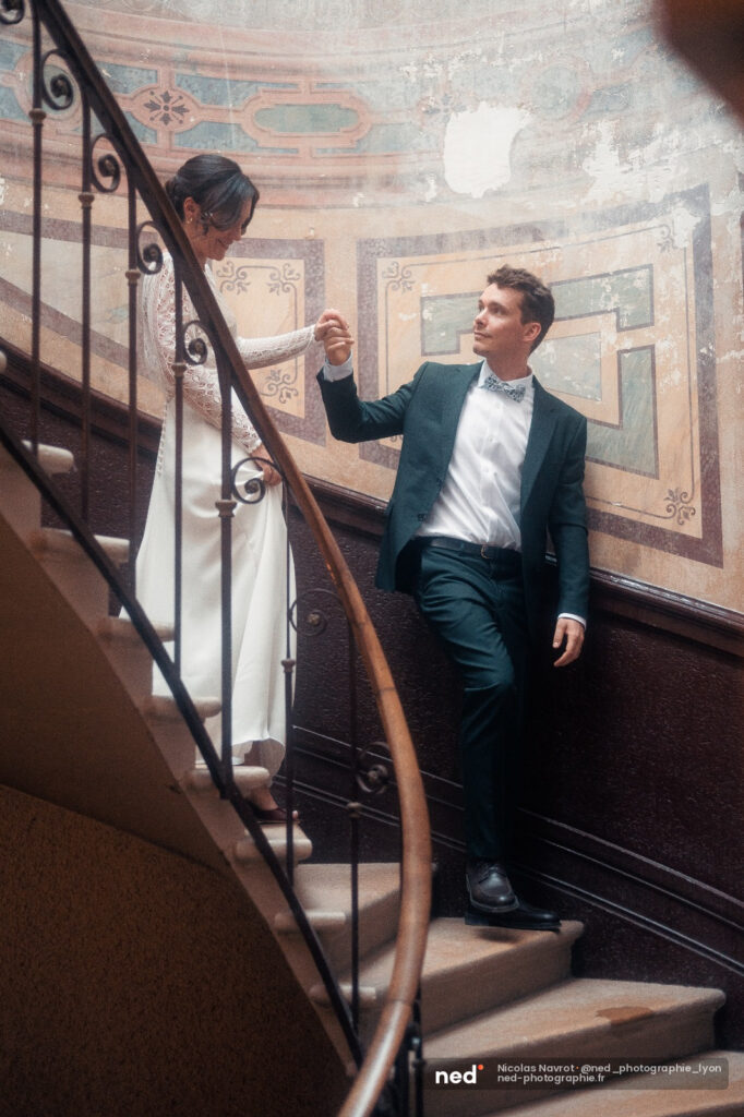 Mariée en robe blanche et marié en costume bleu tiennent leurs mains sur un escalier en colimaçon aux murs peints de motifs pastels