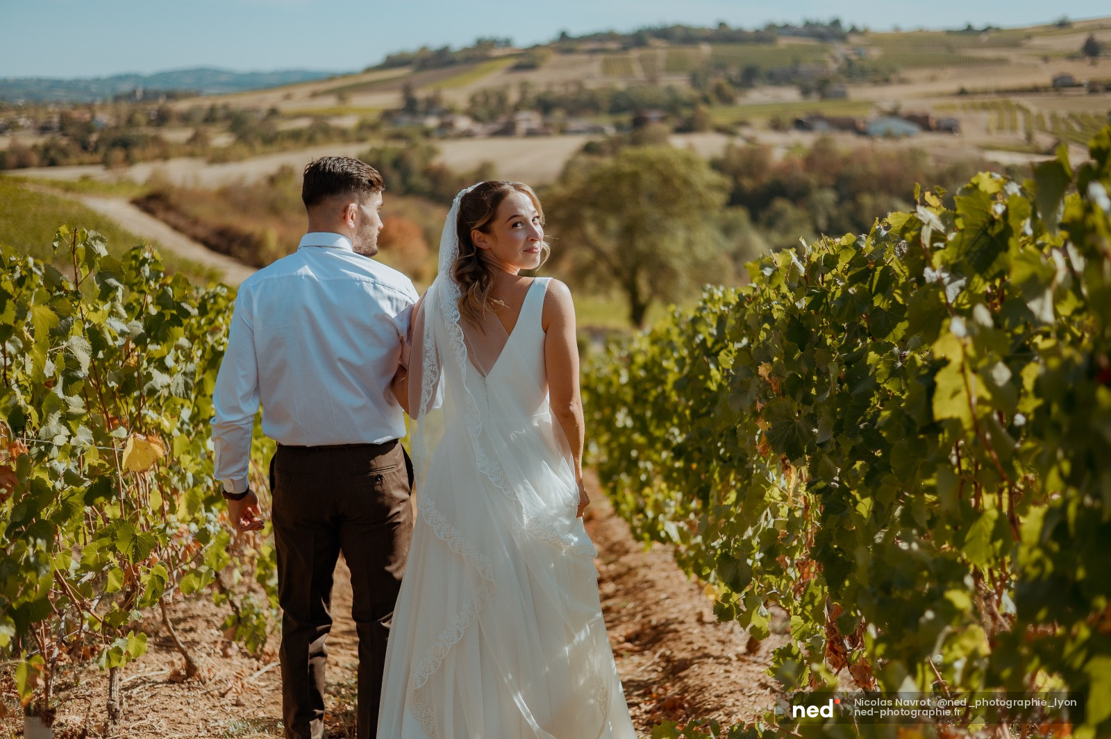Mariée en robe blanche et marié en chemise bleue marchent dans les rangs de vignes, paysage de collines en arrière-plan