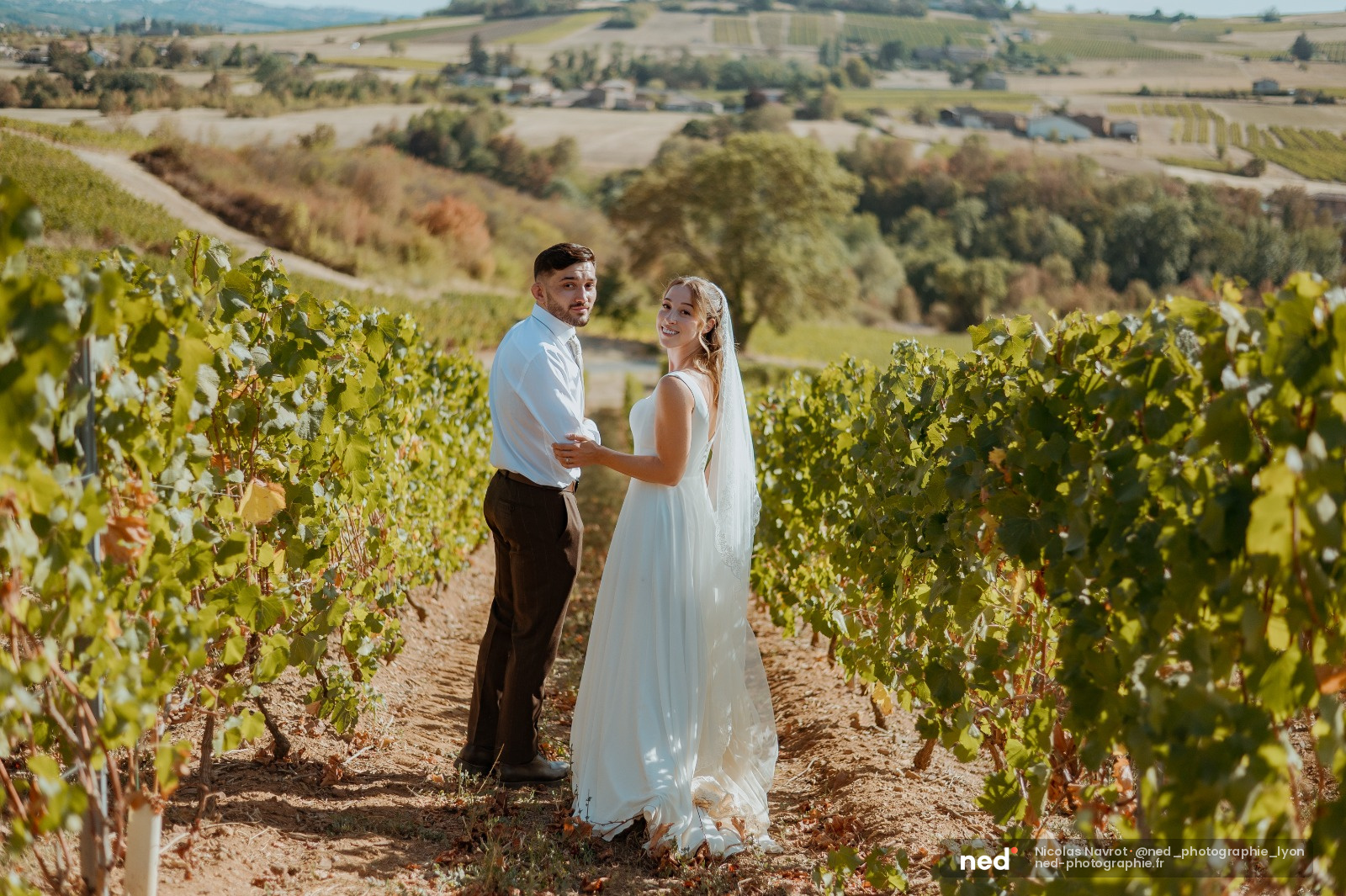 Mariée en robe blanche et marié en chemise bleue posent entre les rangées de vignes, vallée du Rhône en arrière-plan