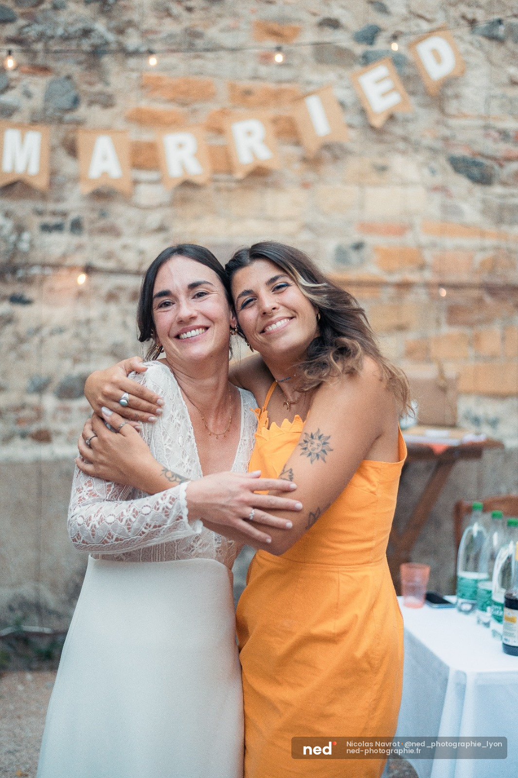 Deux femmes en robe de mariée et robe orange souriantes, enlacées devant un vieux mur en briques avec une guirlande MARRIED