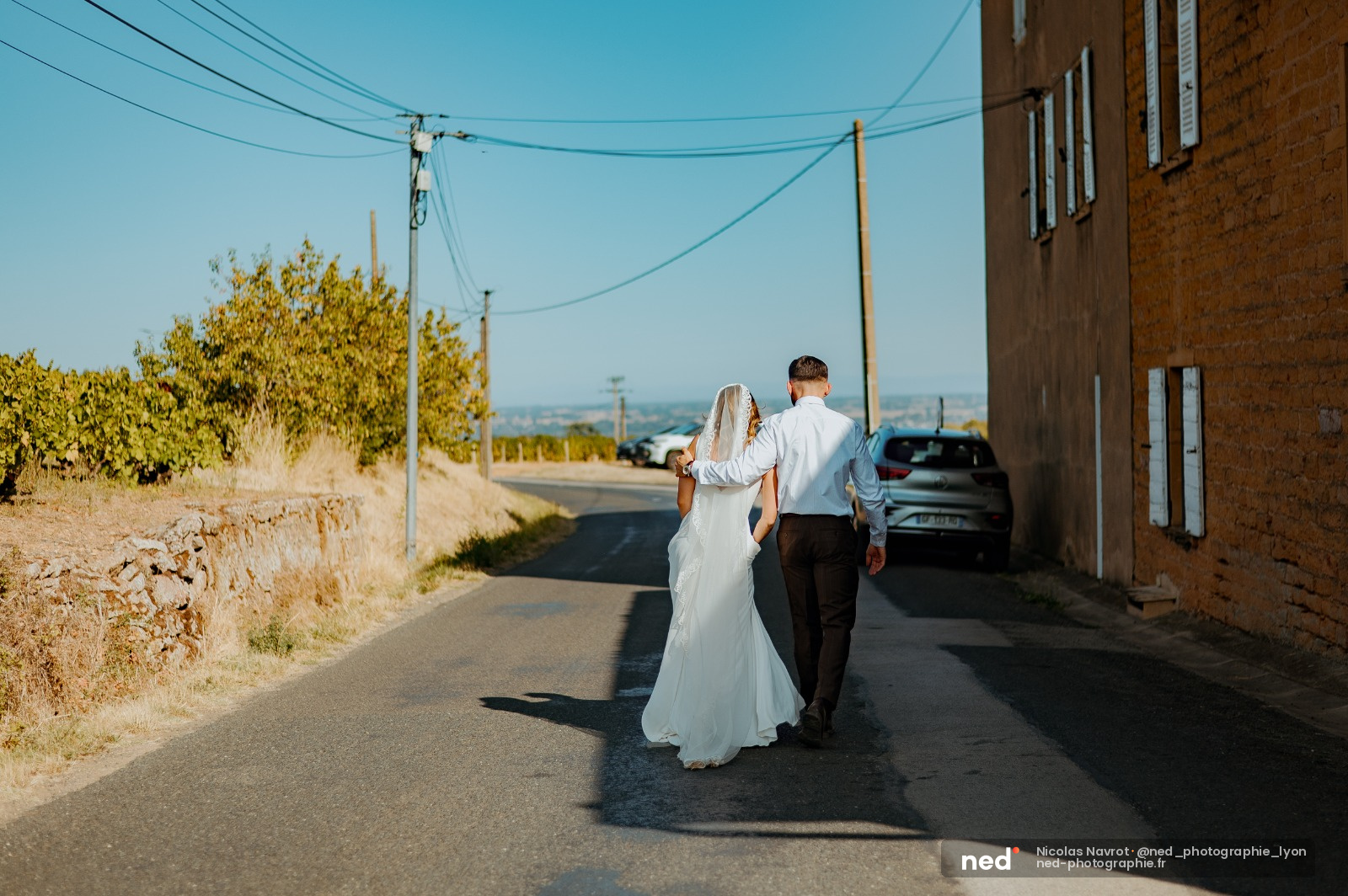 Mariée en robe blanche et voile marchant aux côtés du marié en chemise blanche sur un chemin de campagne, ombres nettes au sol, ciel bleu dégagé.