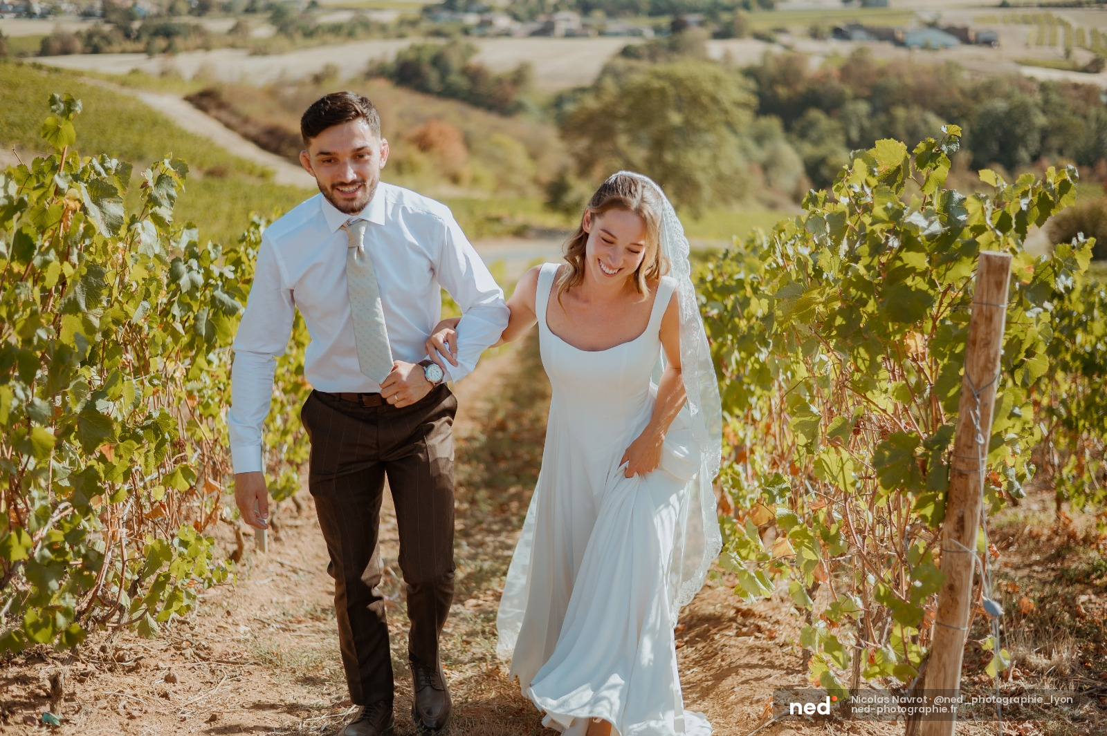 Mariés courant dans les rangs de vignes, elle en robe blanche avec voile, lui en chemise bleue, paysage vallonné en arrière-plan