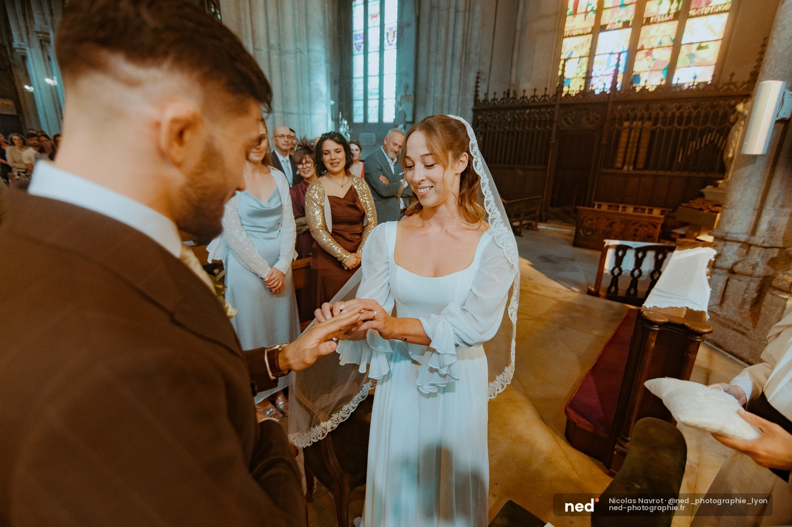 Mariée en robe blanche et voile souriant en échangeant les alliances avec le marié dans une église, invités en arrière-plan