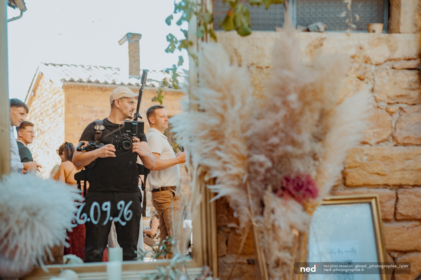 Caméraman en noir avec perche micro filme dans une ruelle pavée aux murs de pierre, entouré de l'équipe de production mariage