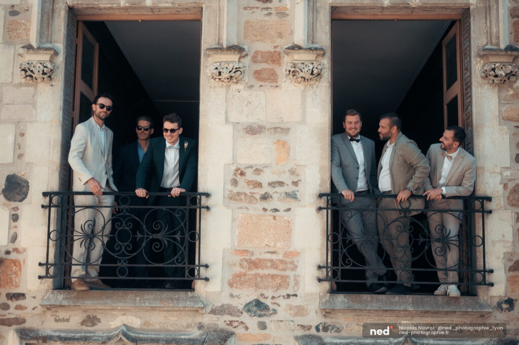 Six hommes en costumes posent sur deux balcons en fer forgé d'une façade en pierre patinée, lumière naturelle.