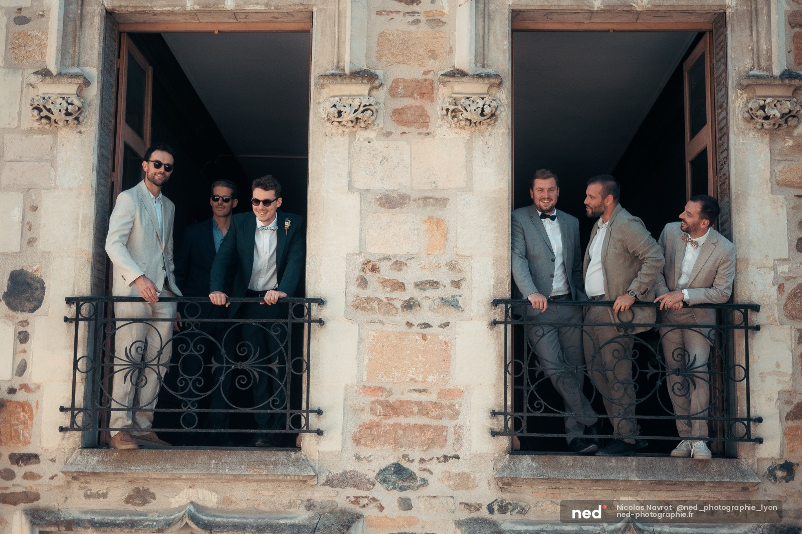 Six hommes en costumes posent sur deux balcons en fer forgé d'une façade en pierre patinée, lumière naturelle.