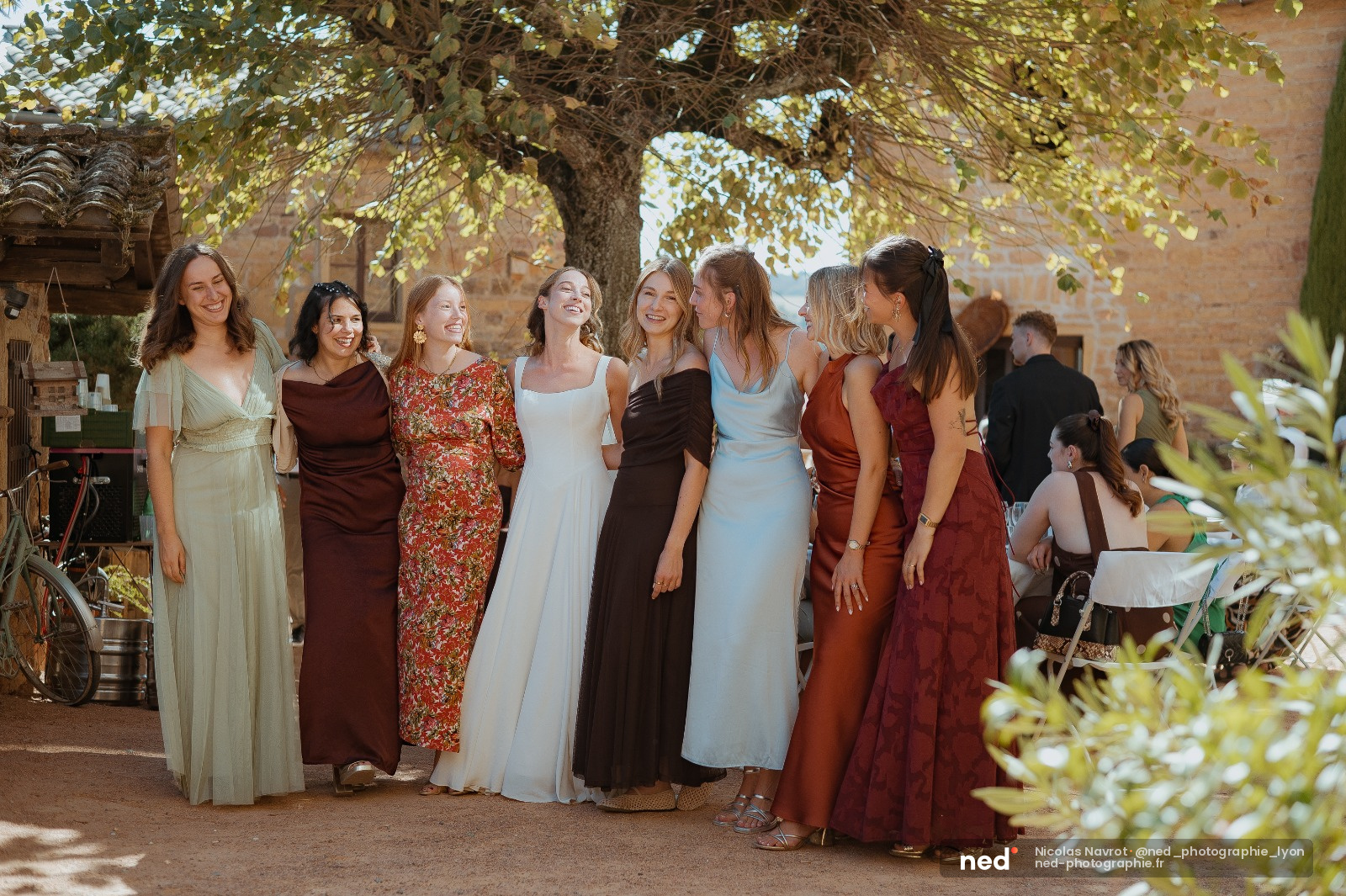 Huit femmes en robes de couleurs variées posent ensemble sous un grand arbre dans une cour en pierre, souriant à la lumière du jour