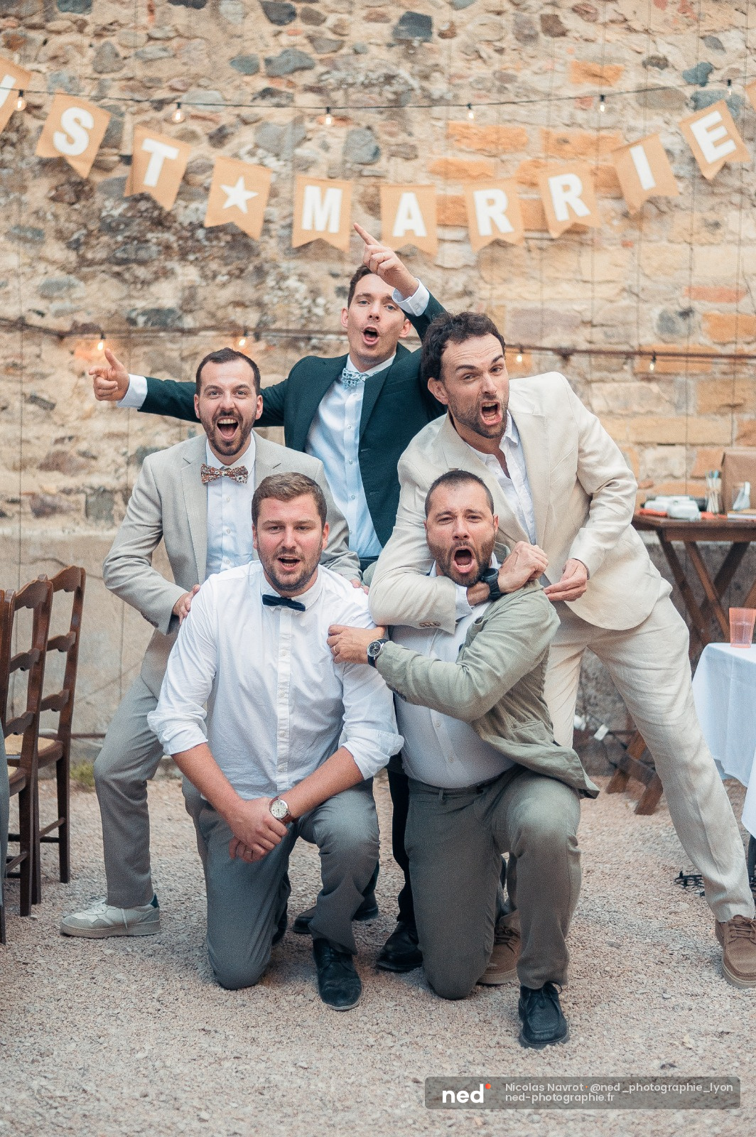 Cinq hommes en costumes posent devant un mur de pierre brute, souriants et énergiques, avec une banderole Just Married accrochée au-dessus