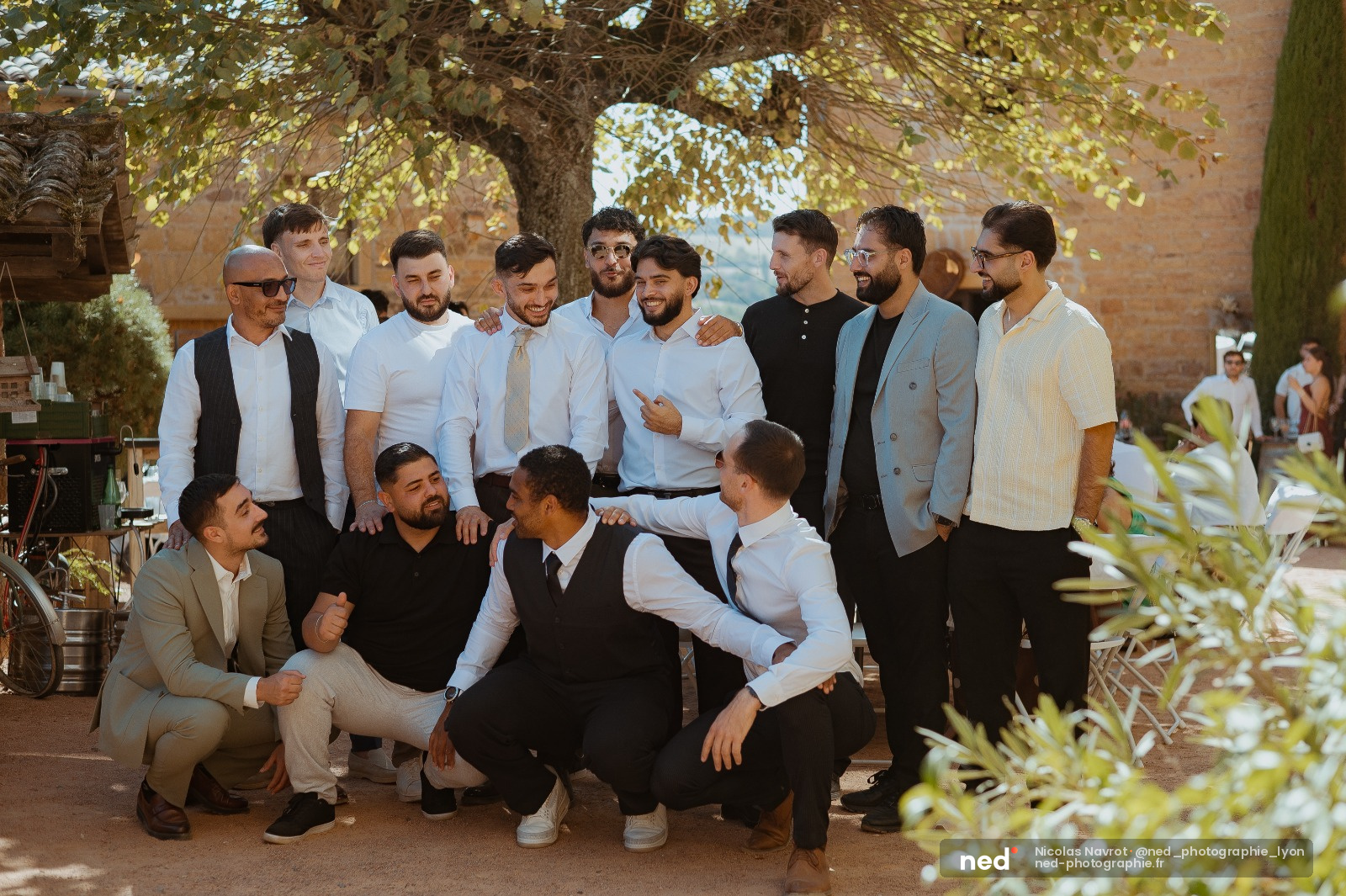 Groupe de douze hommes en tenues de mariage posent ensemble sous un arbre dans une cour, riant et complices