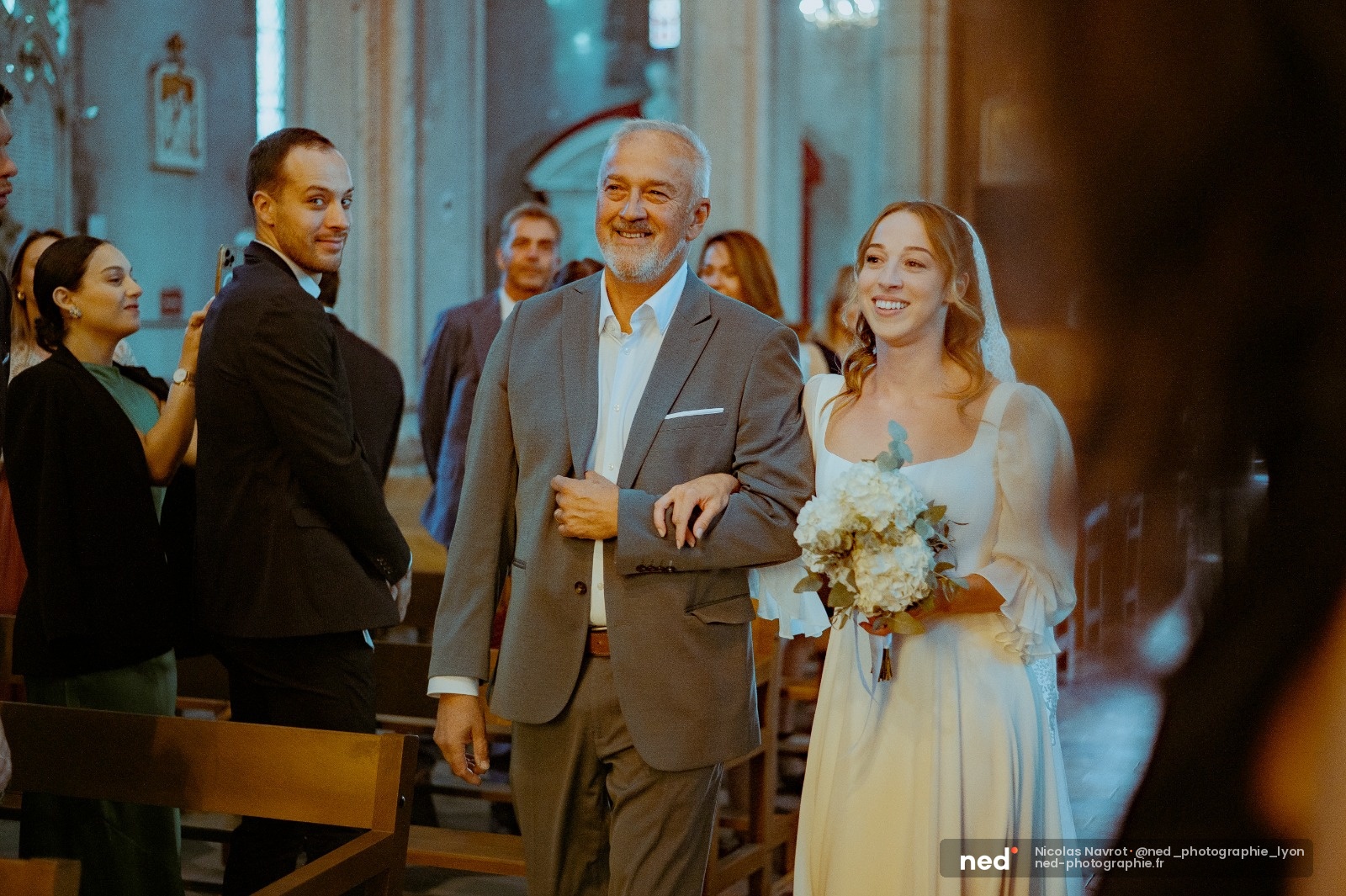 Père en costume gris accompagnant sa fille en robe blanche dans l'allée d'une église, invités souriants en arrière-plan