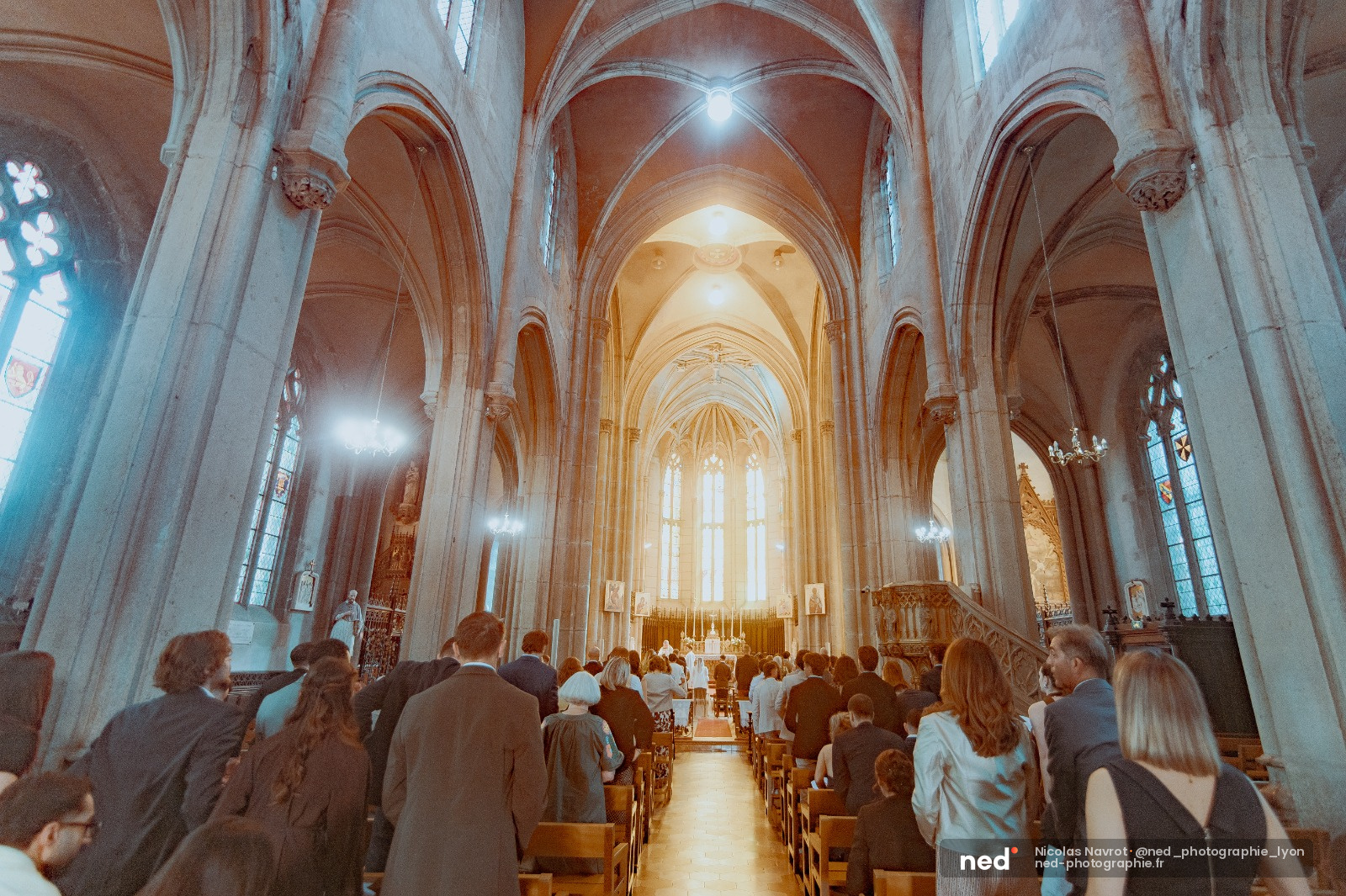 Invités de mariage assis dans les bancs d'une cathédrale gothique, lumière dorée traversant les vitraux