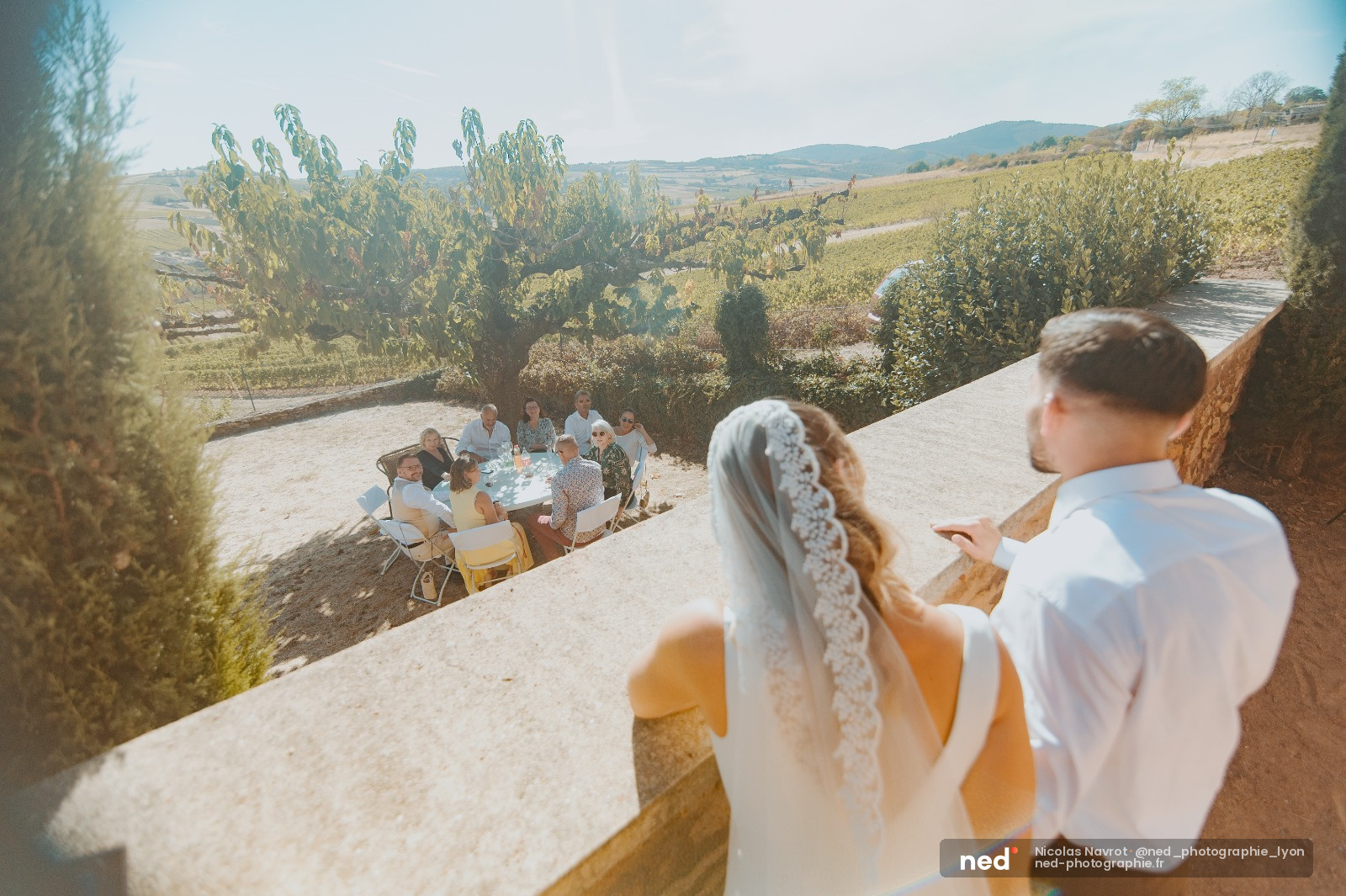 Mariée en robe blanche et marié regardant leurs invités assis à table dans un jardin toscan avec collines en arrière-plan