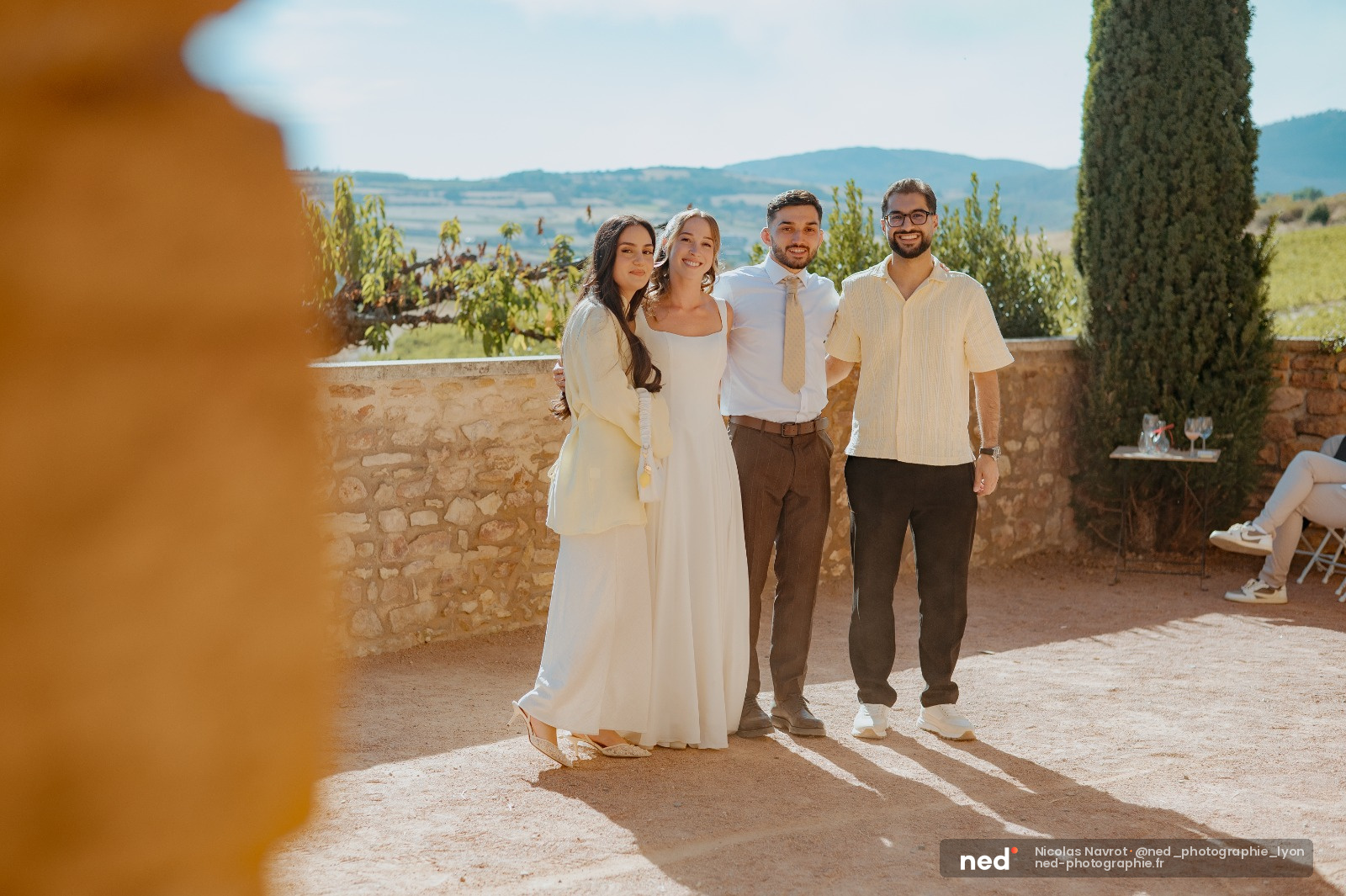 Couple de mariés avec deux témoins posent souriant devant un paysage de collines toscanes, pierre ocre en premier plan