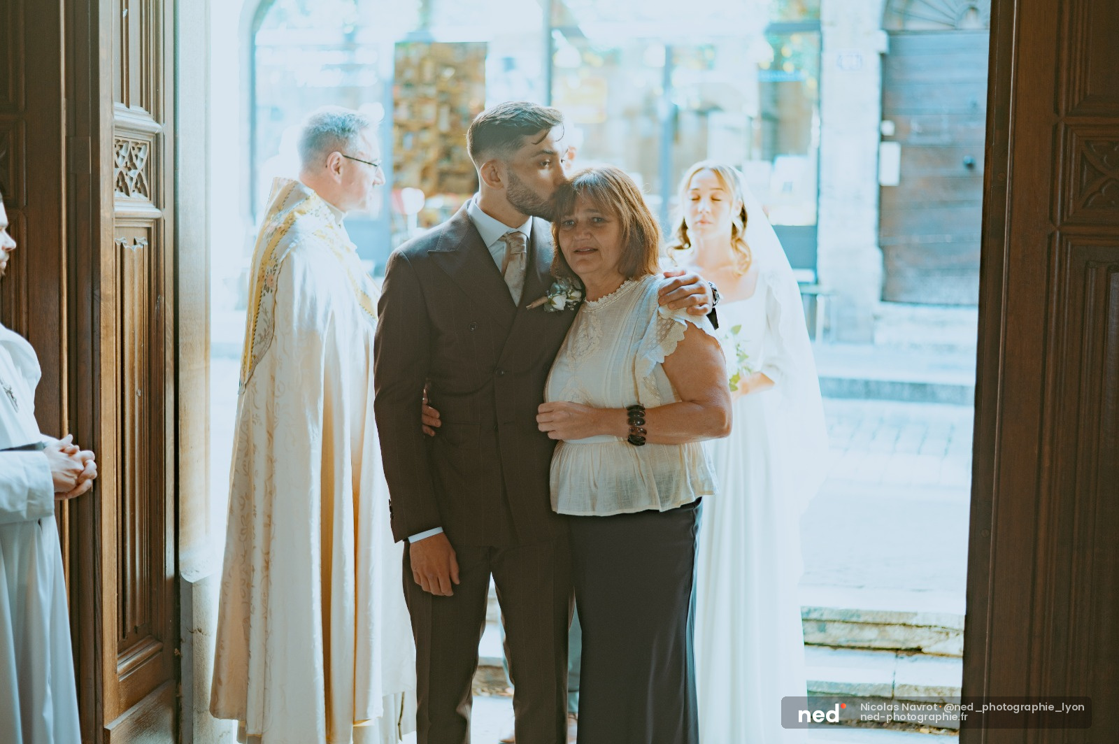 Marié en costume gris embrasse sa mère vêtue de blanc et bleu ciel dans une église lumineuse, pendant la cérémonie de mariage