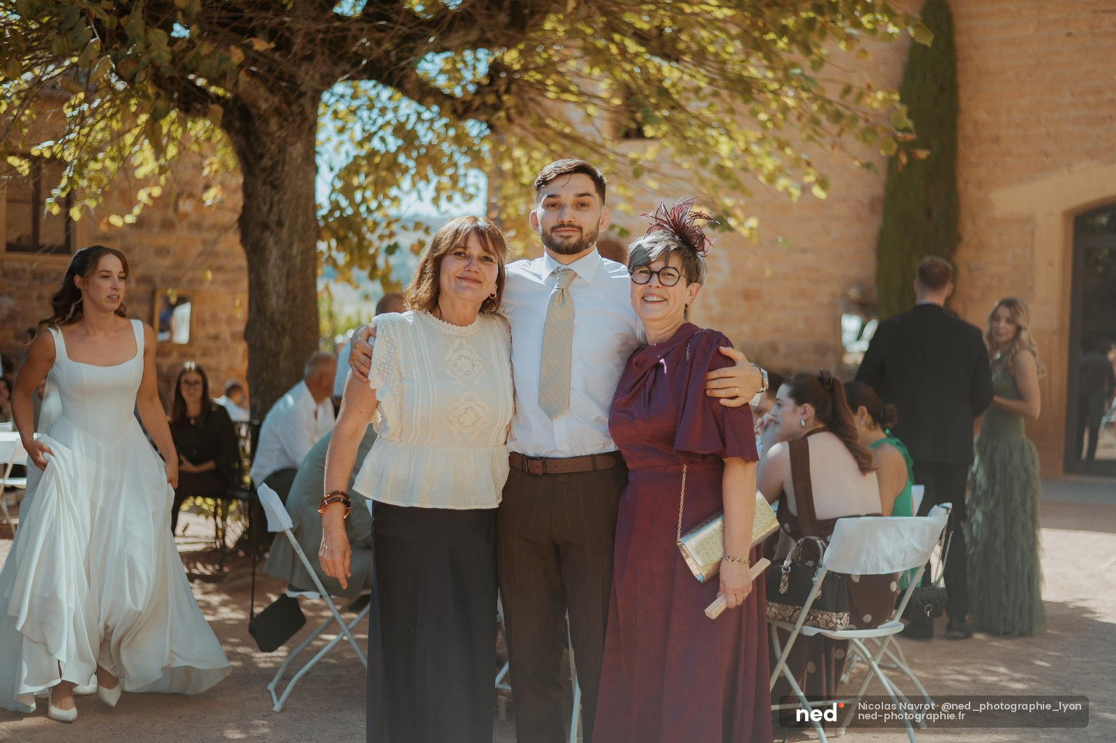 Marié en chemise blanche et cravate pose avec sa mère et sa grand-mère sous un grand arbre, lumière de jour douce