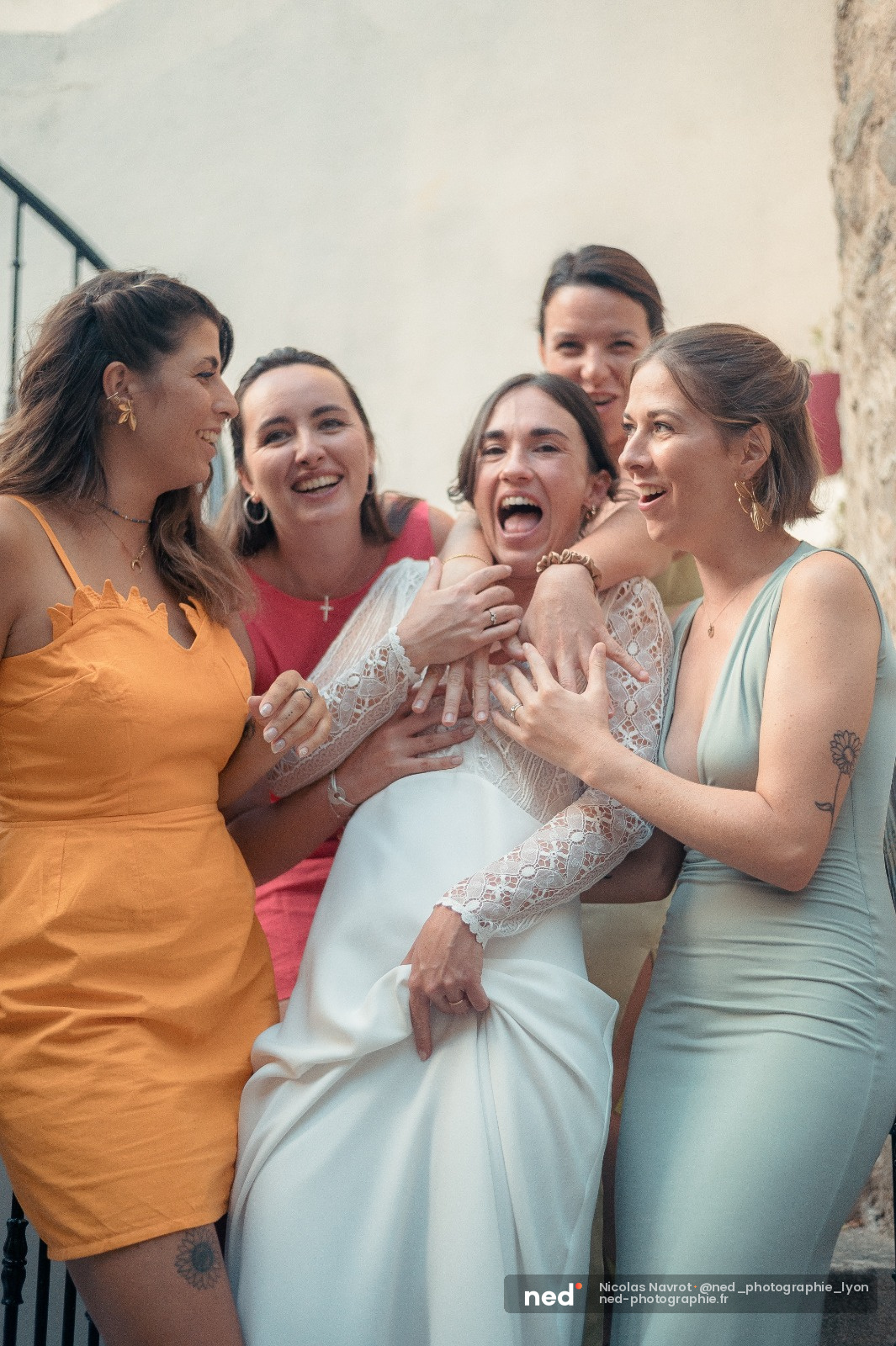 Mariée en robe blanche dentelle entourée de cinq femmes en robes colorées, toutes riant ensemble devant un mur en pierre