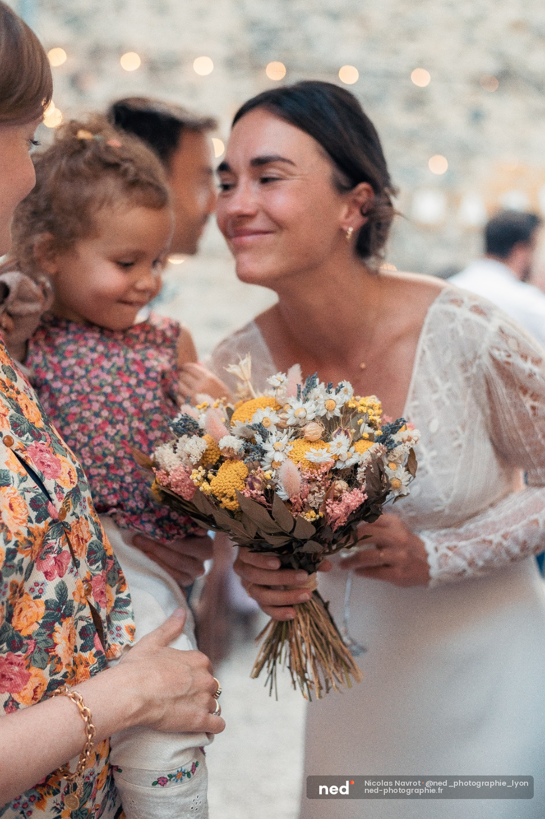 Mariée en robe dentelle blanche tient son bouquet de fleurs sèches, sourit à une petite fille en robe fleurie à ses côtés