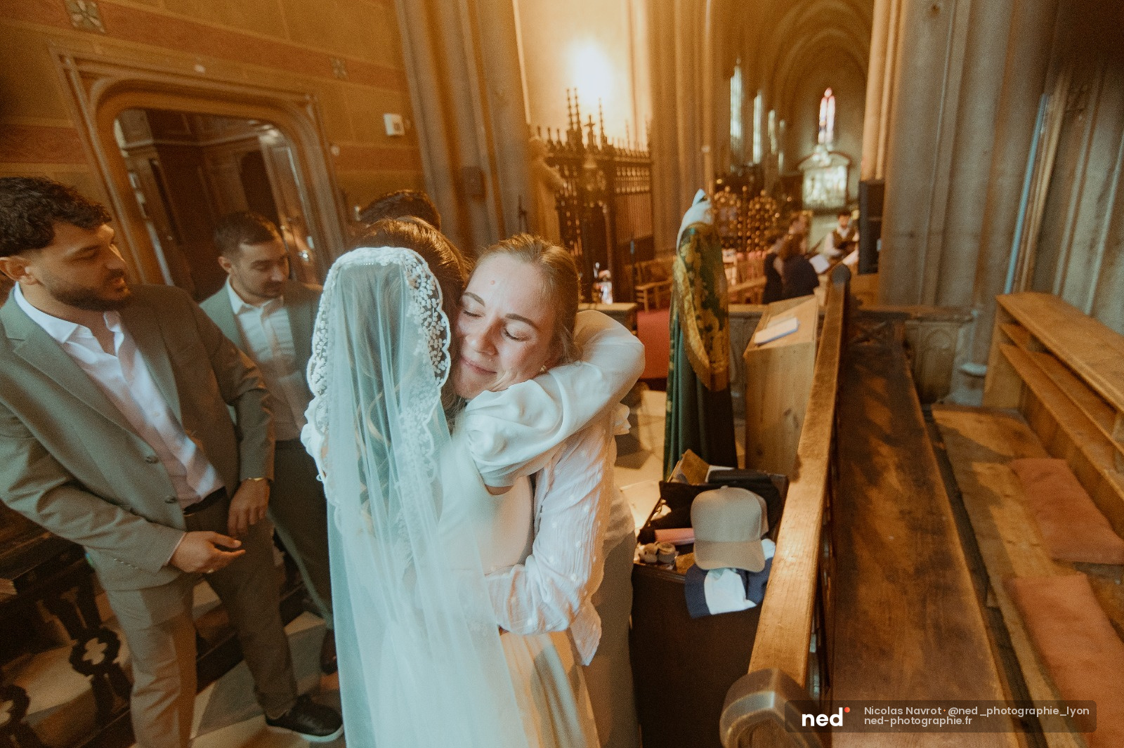 Mariée en robe blanche et voile enlacée par un proche dans la nef d'une église en bois, invités en arrière-plan