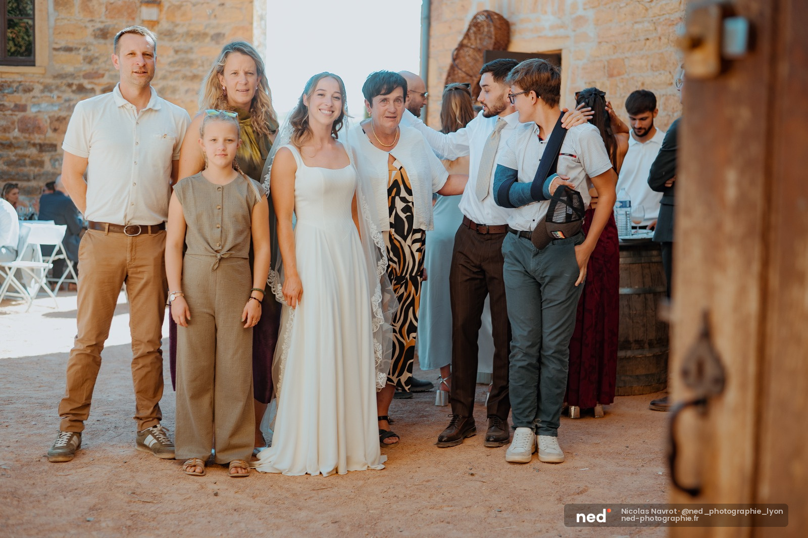 Mariée en robe blanche au centre entourée de famille et amis en tenues colorées dans une cour en pierre, lumière naturelle douce