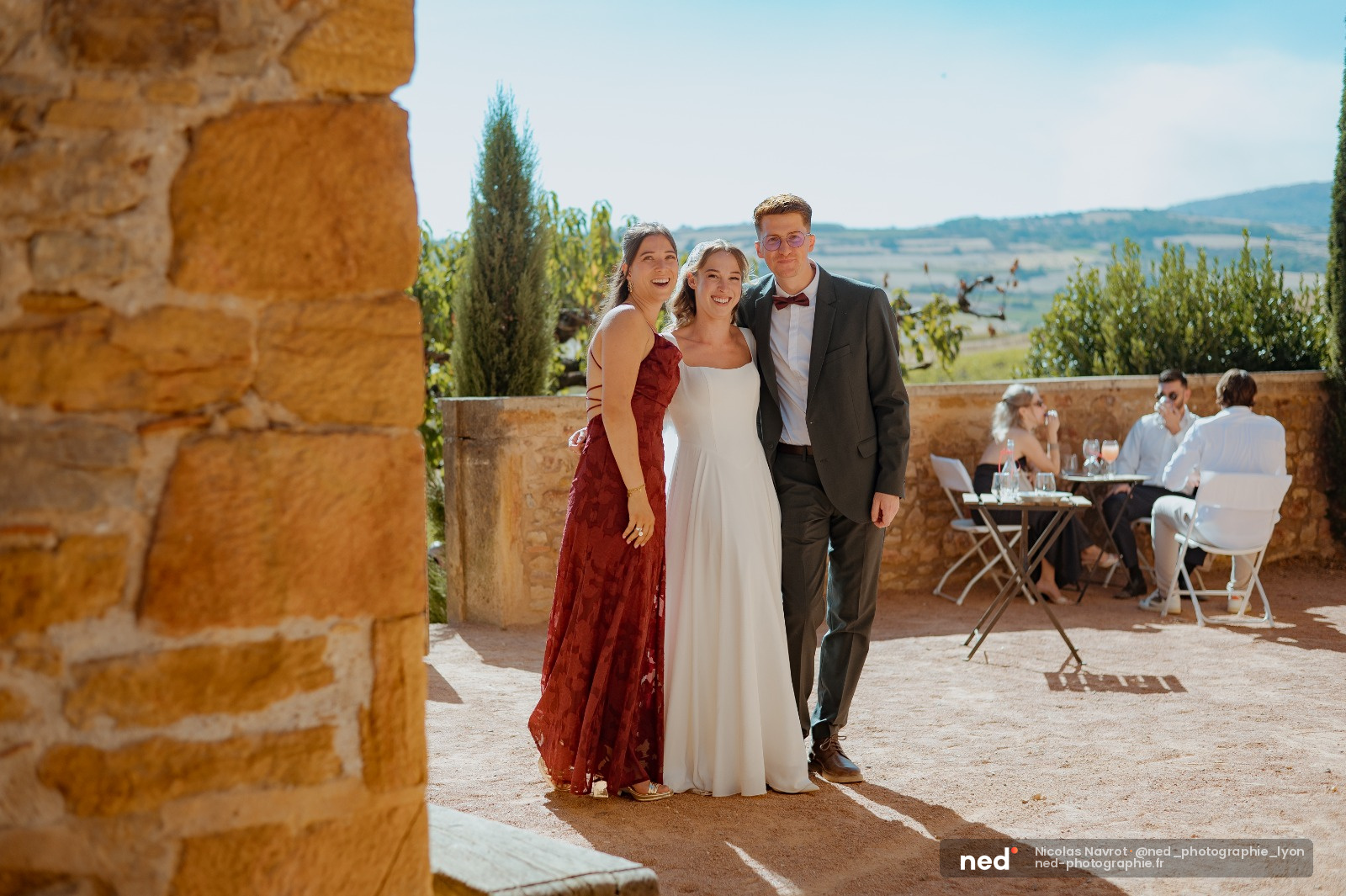 Mariée en robe blanche avec deux invités souriants sur une terrasse en pierre avec vue sur la campagne toscane