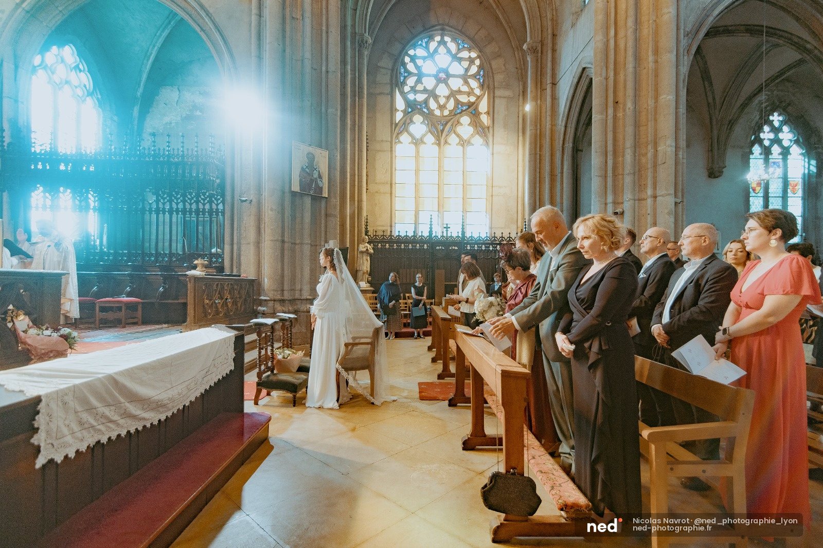 Mariée en robe blanche devant l'autel d'une cathédrale gothique, invités assis dans les bancs, lumière naturelle par les vitraux