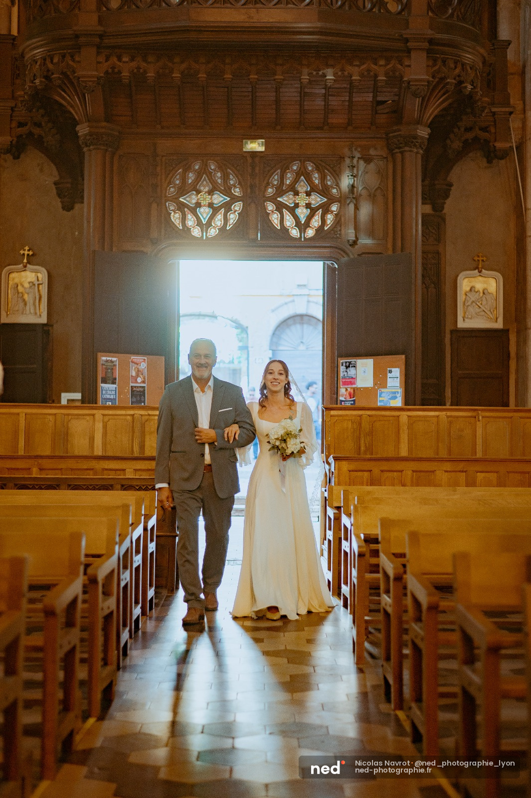 Père en costume gris et mariée en robe blanche remontent l'allée d'une église en bois, lumière naturelle par l'entrée