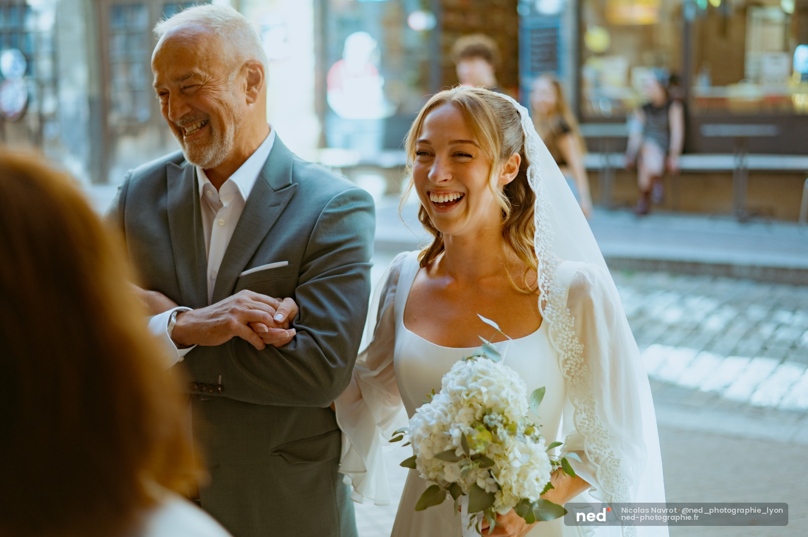 Mariée en robe blanche et voile souriante, tenant un bouquet de fleurs, marchant bras dessus bras dessous avec son père en costume gris dans une rue pavée en extérieur