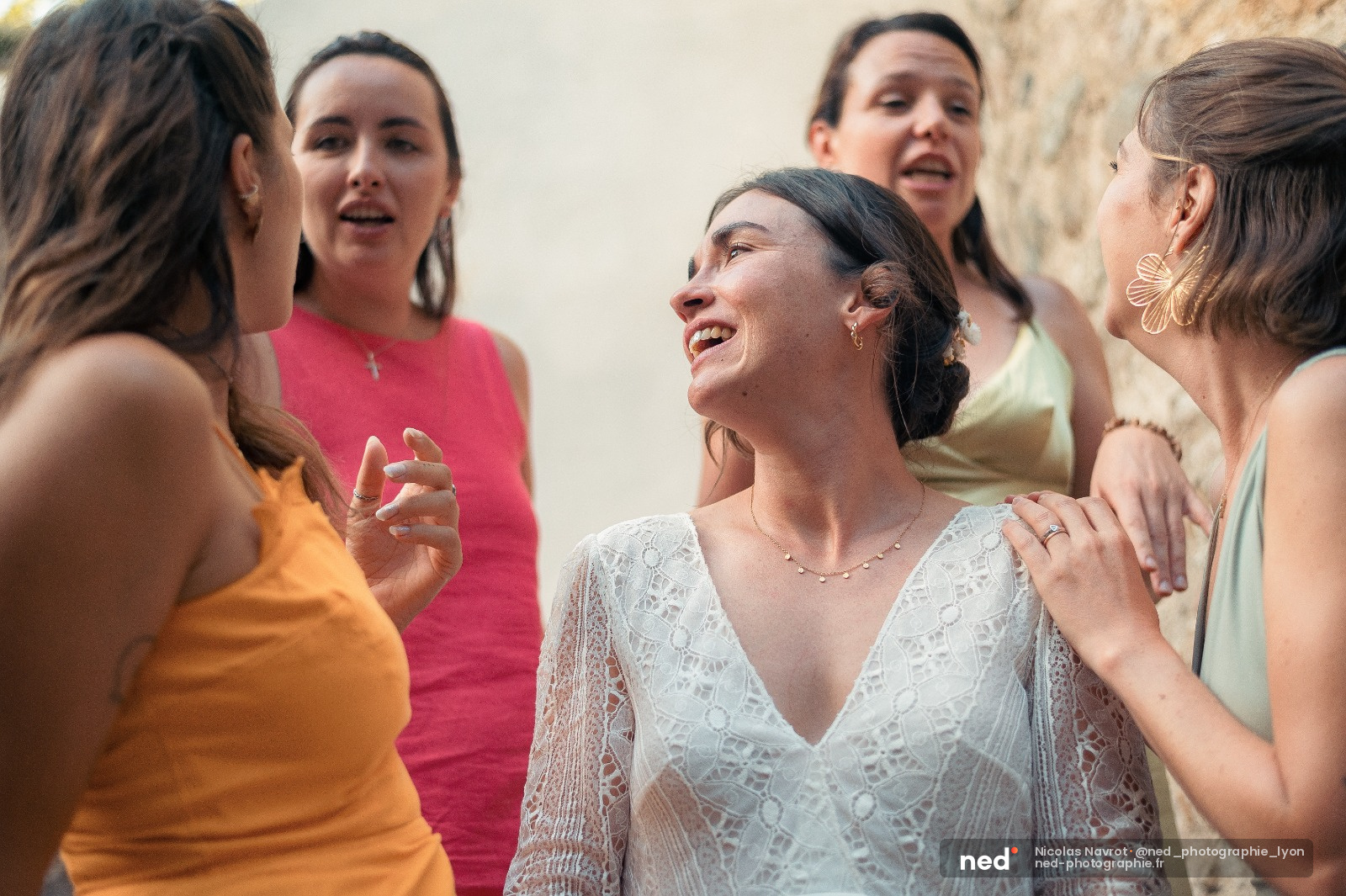 Mariée en robe blanche dentelle riant au milieu de ses demoiselles d'honneur en robes colorées contre un mur de pierre