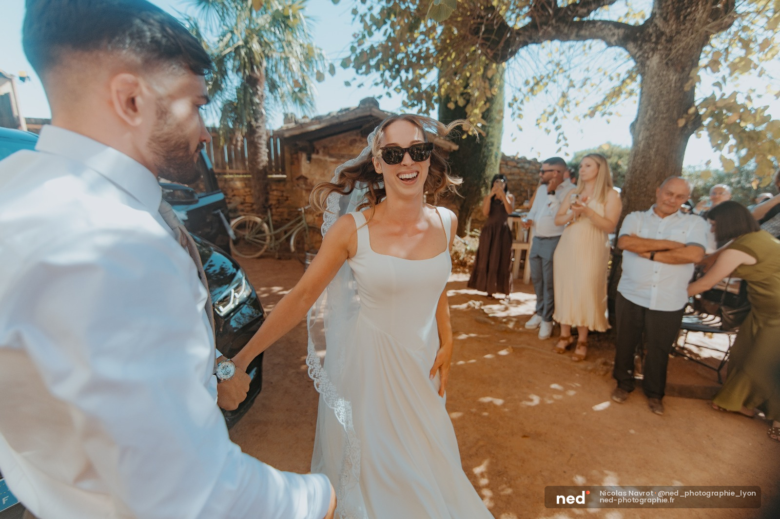 Mariée en robe blanche et lunettes de soleil rit avec le marié en chemise bleue sous un grand arbre, invités en arrière-plan