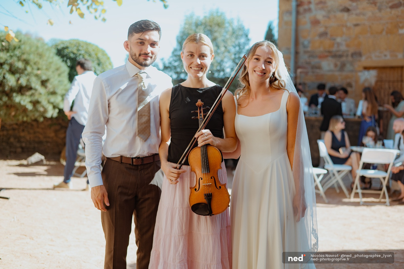 Mariée en robe blanche avec voile, violoniste en robe rose tenant son instrument, et témoin en chemise blanche posent devant une bâtisse en pierre lors d'un mariage en extérieur