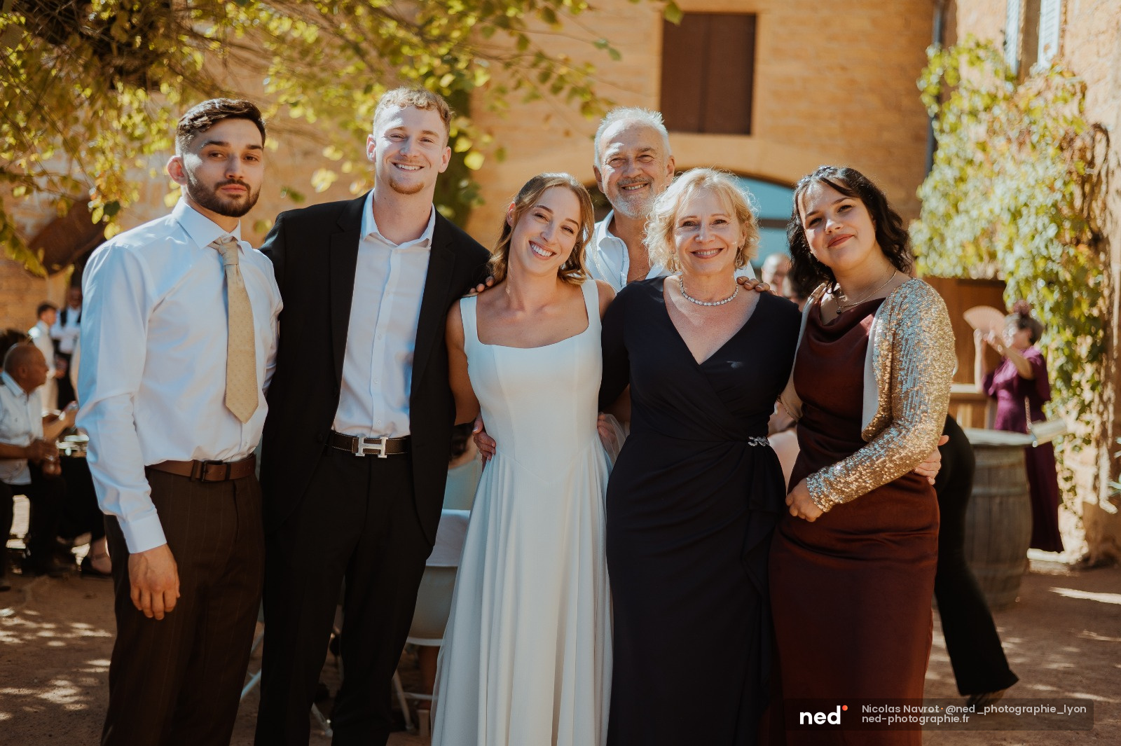 Mariée en robe blanche posant avec sa famille devant une facade en pierre, lierre vert, lumière du jour