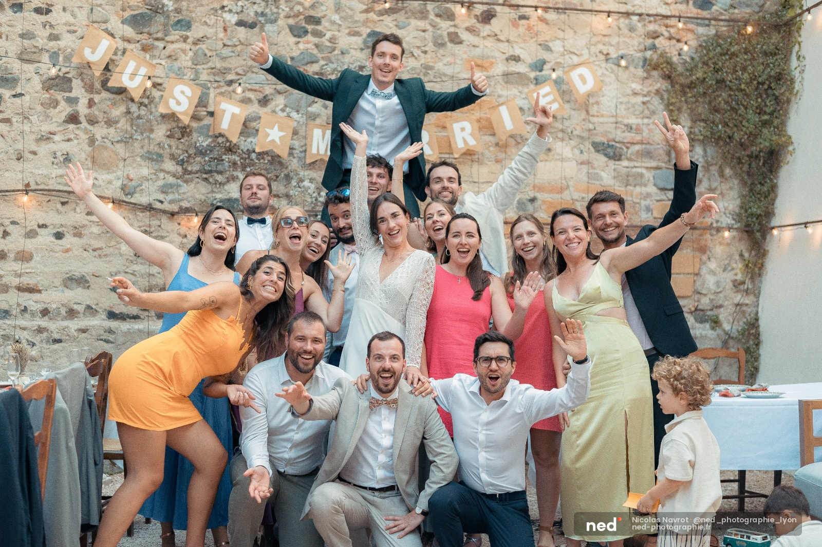Groupe de vingt personnes en tenues colorees pose devant un mur de pierre ancien, bras leves, souriant, avec une banderole just married