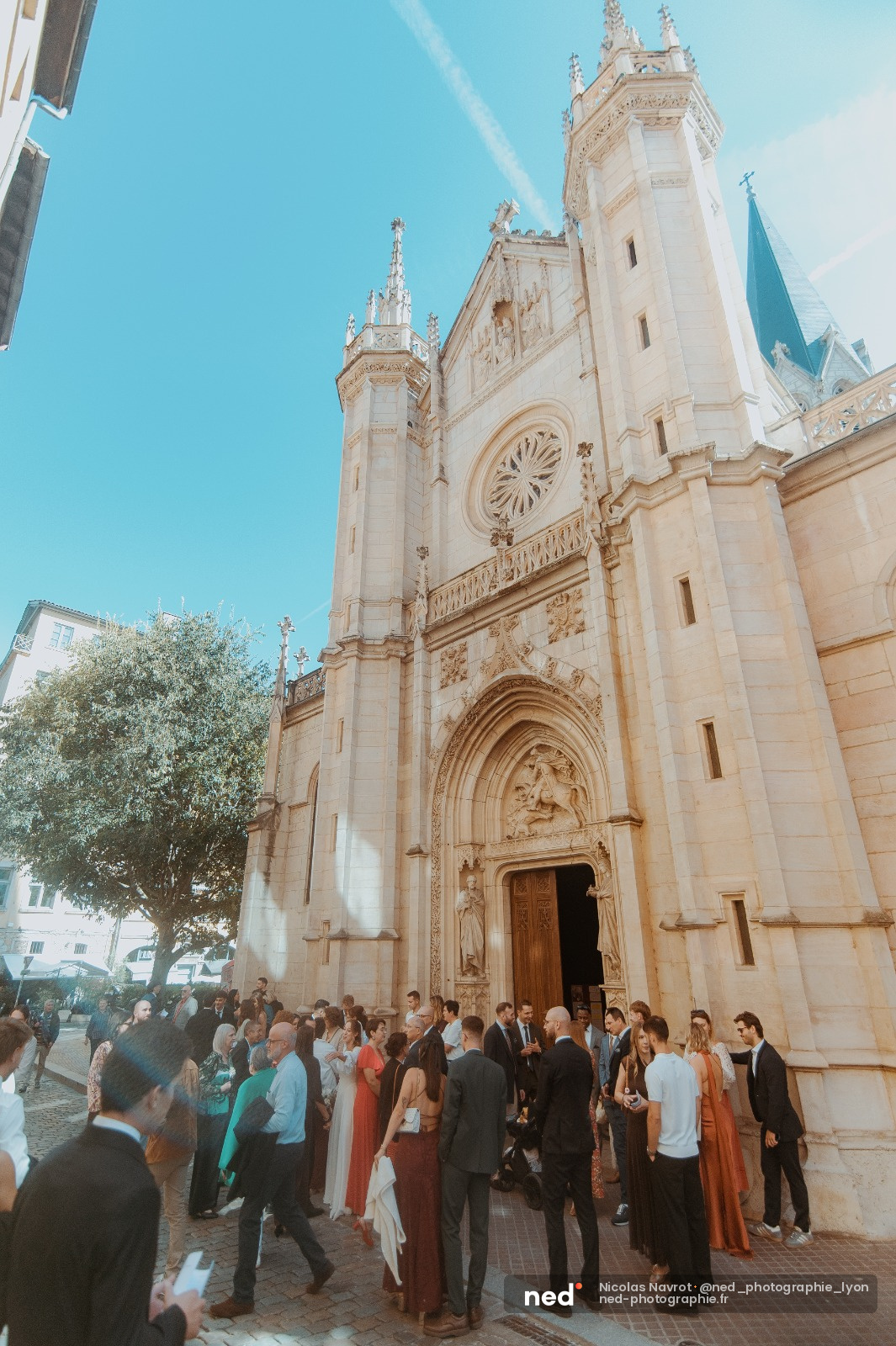 Invités en tenue de mariage rassemblés devant la façade blanche de la basilique Saint-Fourvière, ciel bleu, lumière de jour éclatante