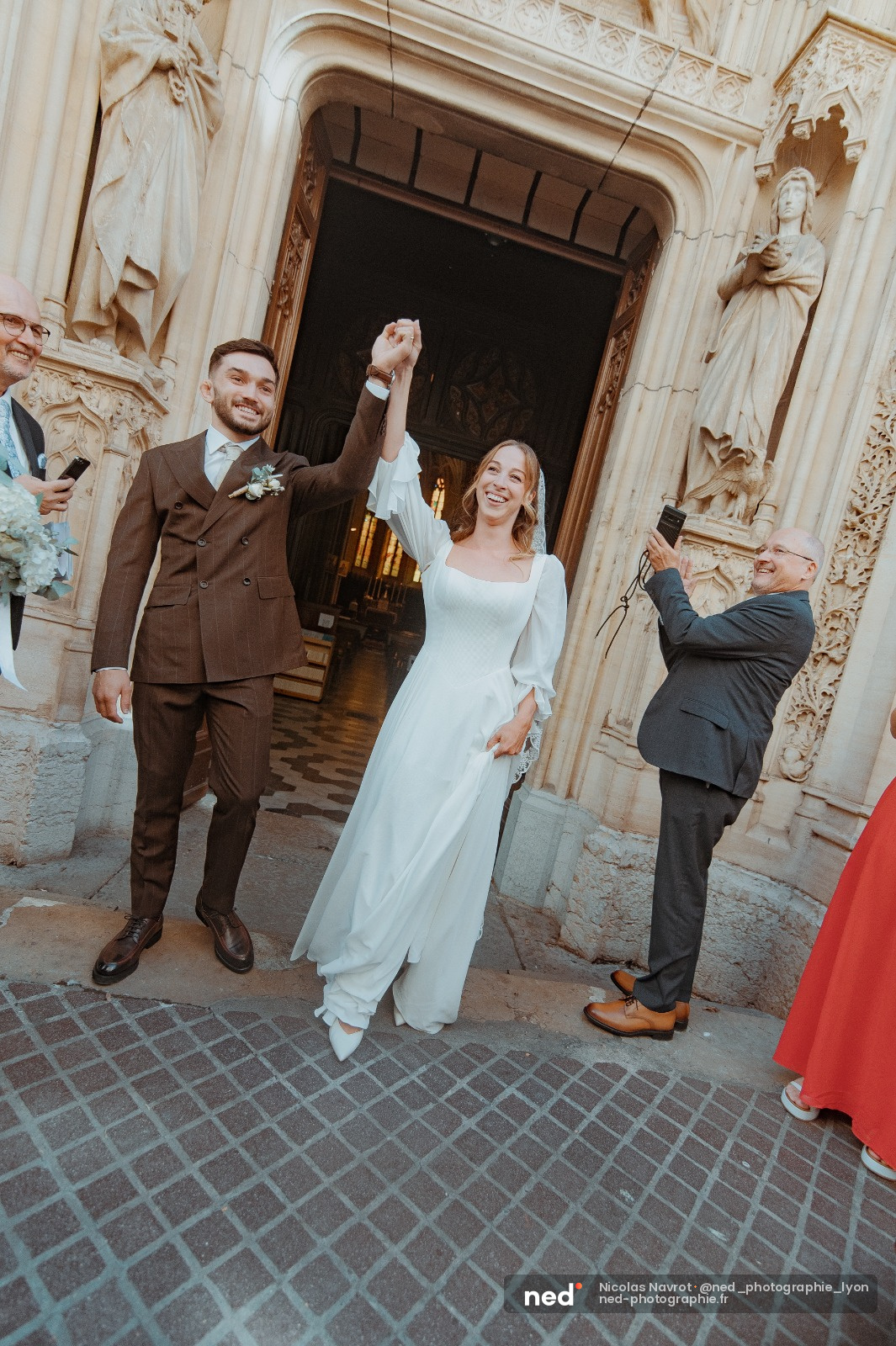Mariee en robe blanche et marie en costume marron levent les mains en souriant devant une vieille eglise en pierre, invites qui filmaient a cote