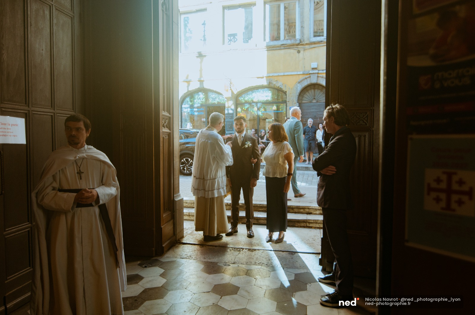 Marié en costume beige discute avec des invités sur le parvis de l'église, prêtre en aube blanche à gauche, bâtiment en pierre jaune en arrière-plan