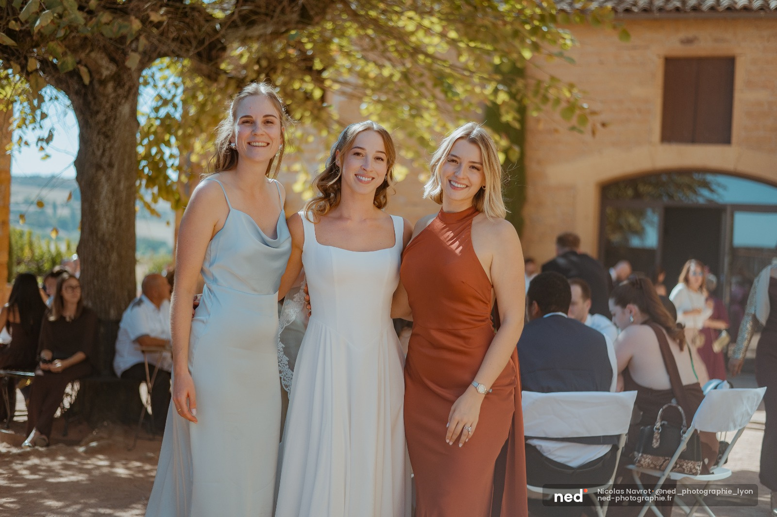 Trois femmes en robes pastel et terracotta sourient face à l'objectif sous un grand arbre, invités de mariage flous en arrière-plan
