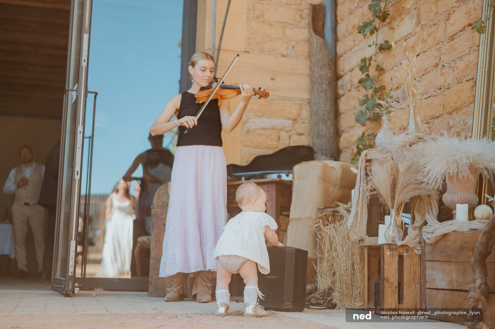 Musicienne en robe rose jouant du violon devant un petit enfant en robe blanche, murs en pierre dorée et ciel bleu