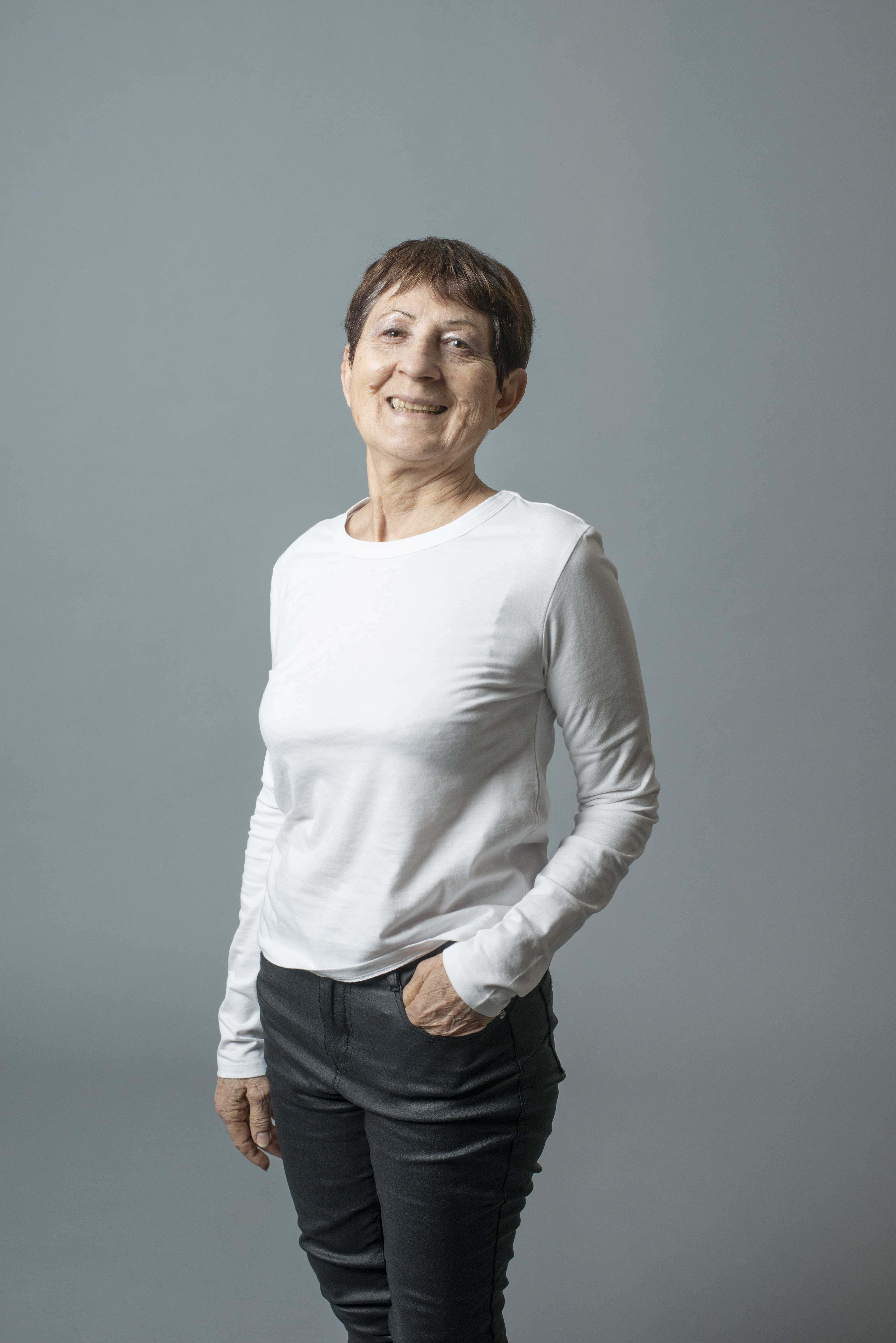 Femme d'âge mûr souriante vêtue d'un pull blanc et pantalon noir, posée devant un fond gris uni
